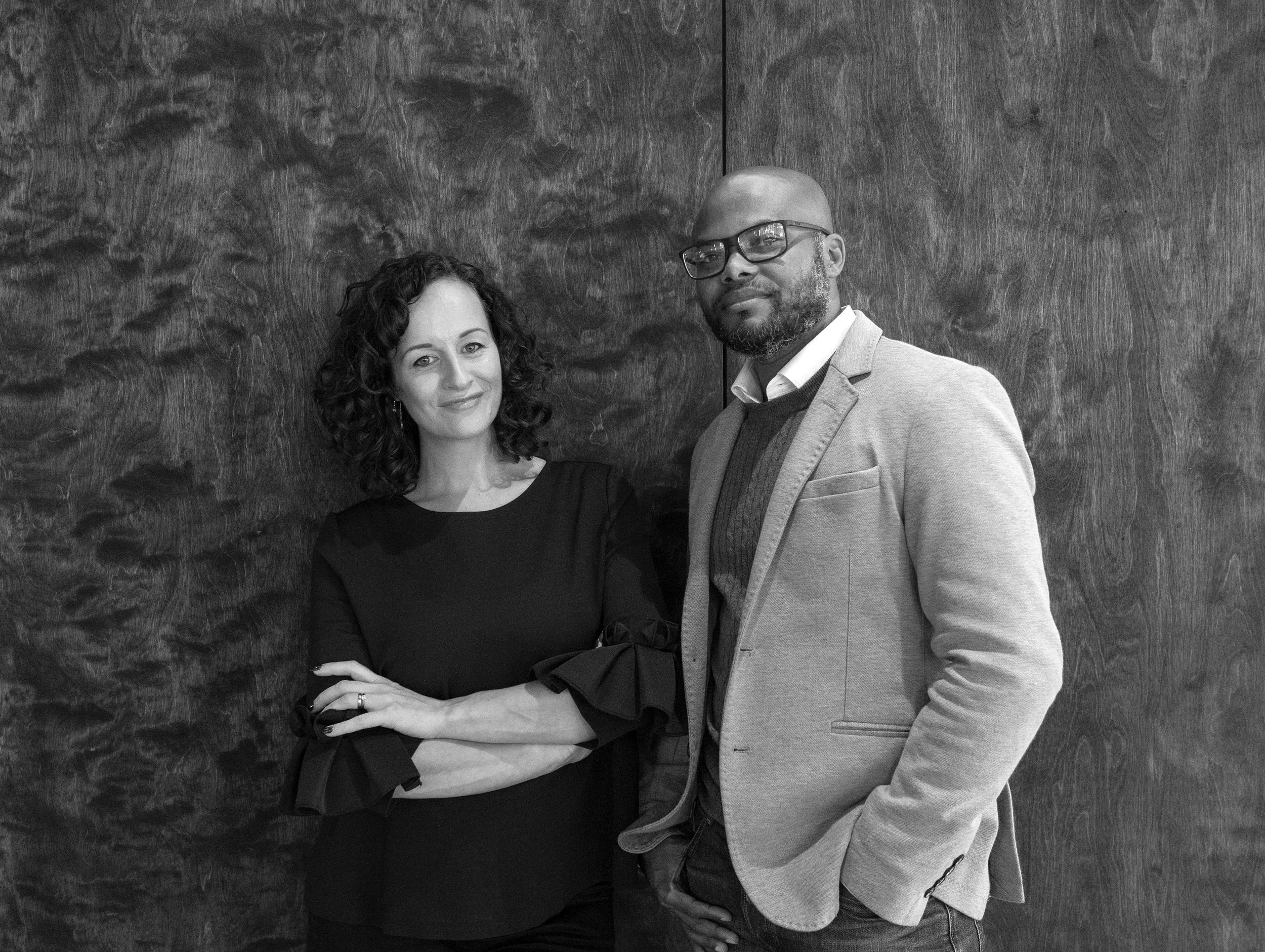 A black and white photo of a woman with curly hair and a man with glasses standing against a textured wall. The woman is smiling with her arms crossed, wearing a dark dress, and the man has one hand in his pocket, wearing a blazer and a sweater.