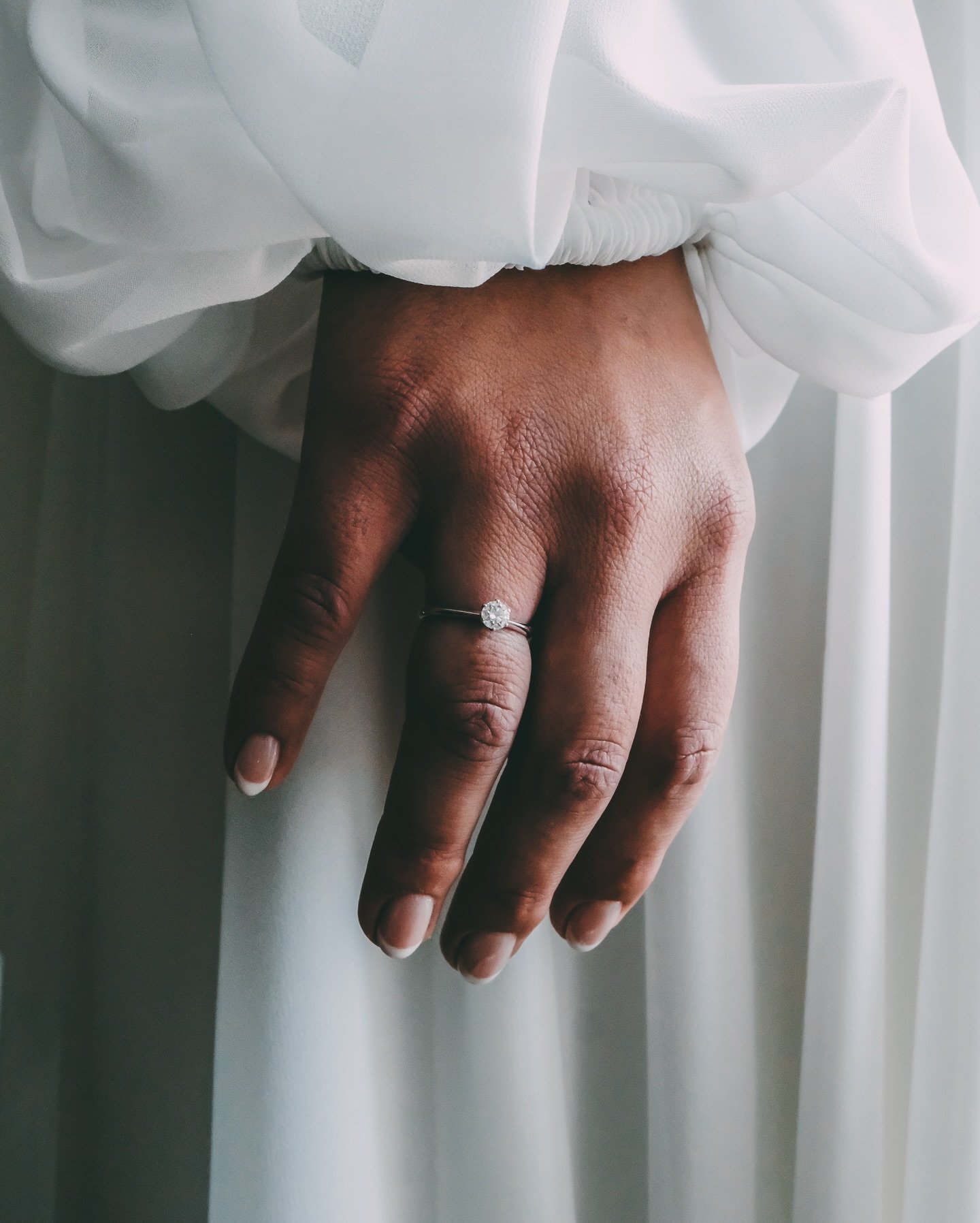 In questo dettaglio ravvicinato, ho voluto fermare il tempo su un punto di luce pura. La mano della sposa, adagiata con naturalezza sul tessuto candido dell&rsquo;abito, diventa il palcoscenico per quel piccolo diamante che racchiude un universo di p
