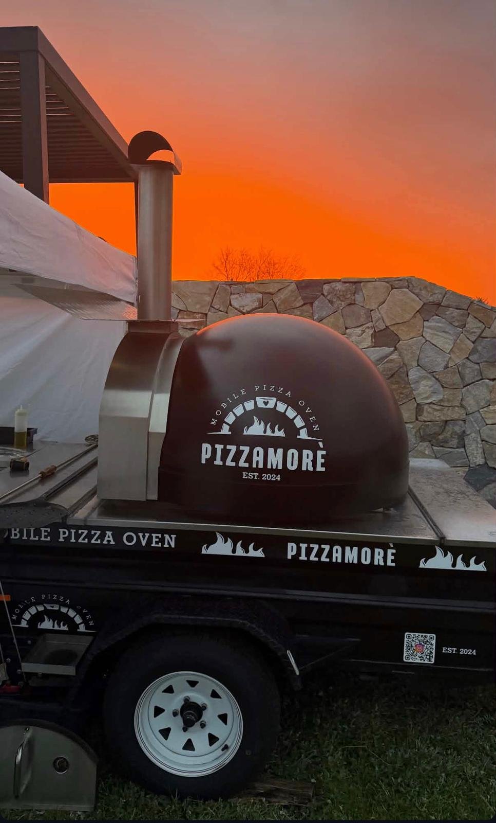 A mobile outdoor pizza oven with a black dome and a logo reading "Pizzamore" against an orange sunset sky. The oven is mounted on a black trailer with branding and a QR code, next to a stone wall.