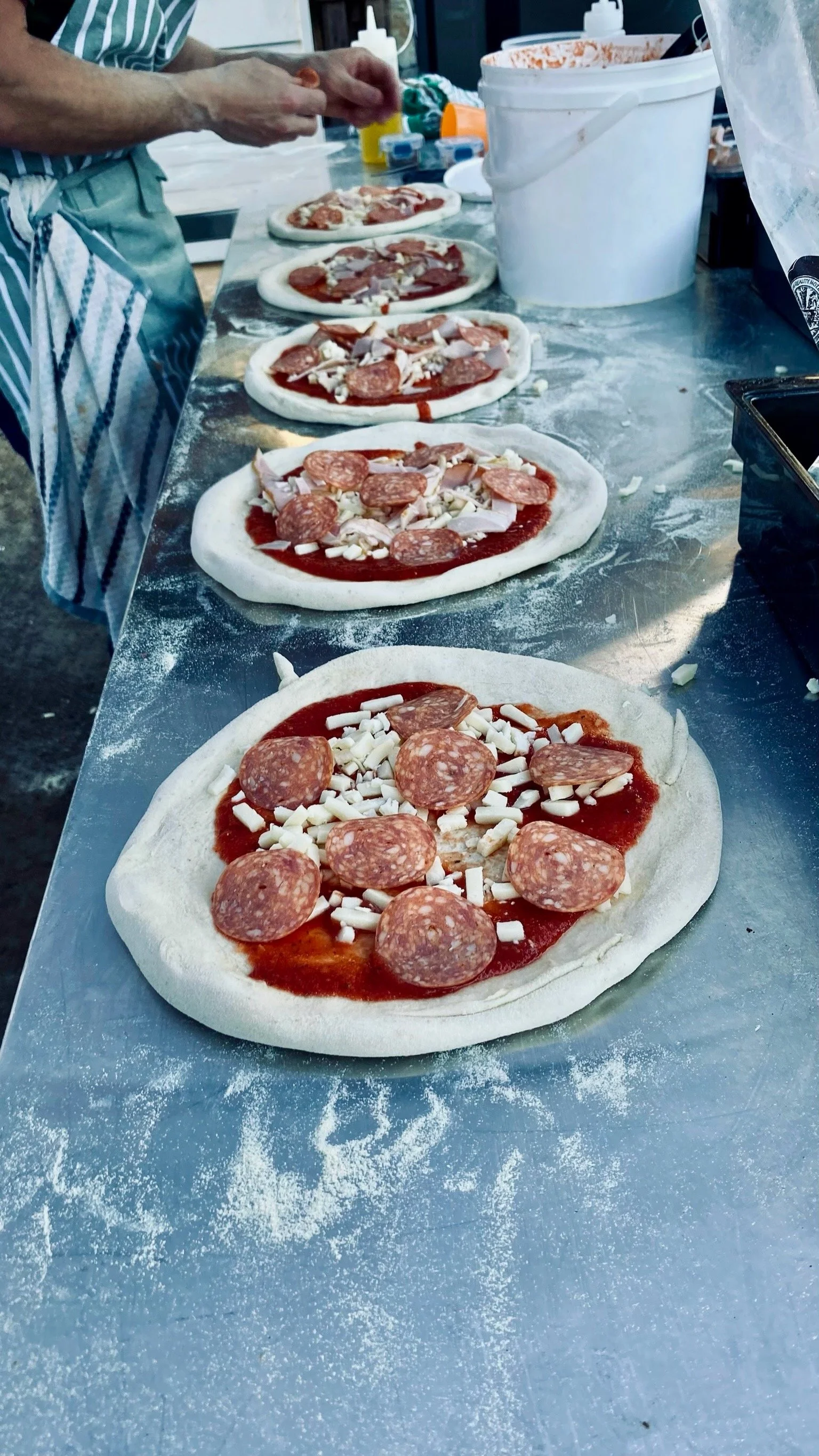 Several unbaked pizzas with pepperoni, ham, and cheese on a floured metal table.