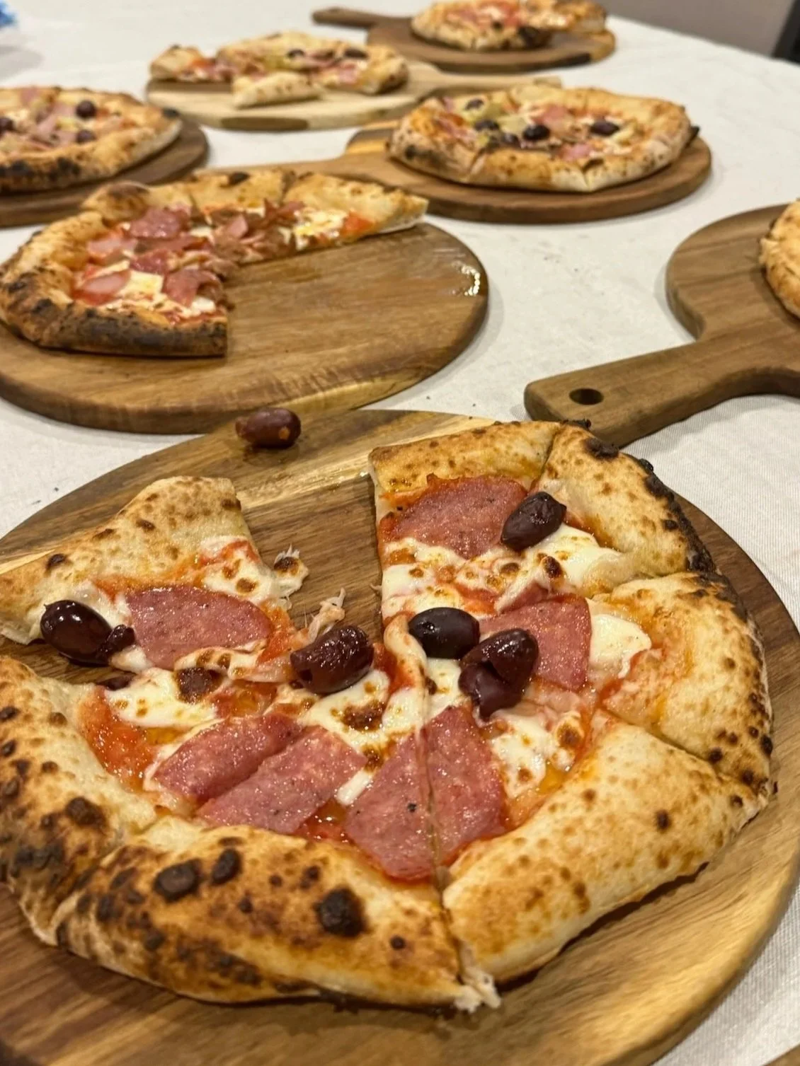 A close-up of a pepperoni and black olive pizza on a wooden serving board, with several other pizzas on similar boards in the background.