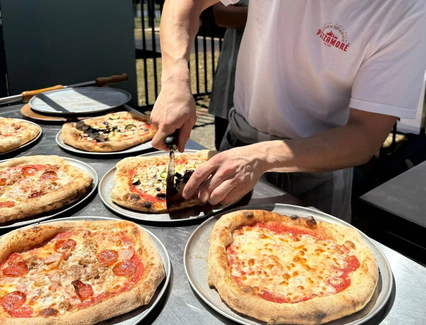 Person slicing a pizza outdoors, with several pizzas on plates on a stainless steel table.