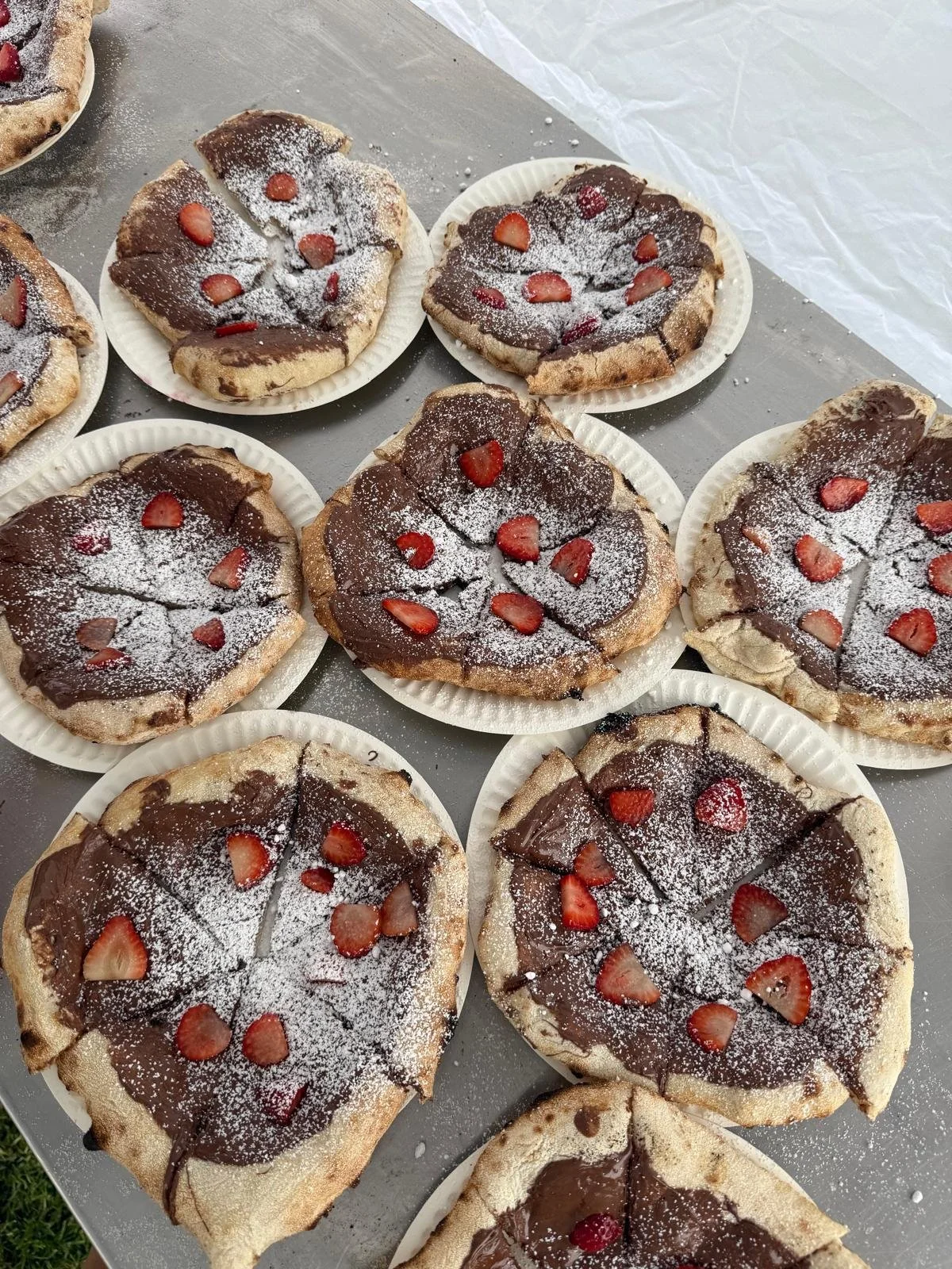Several flatbread pizzas topped with chocolate, sliced strawberries, and powdered sugar, served on white paper plates.
