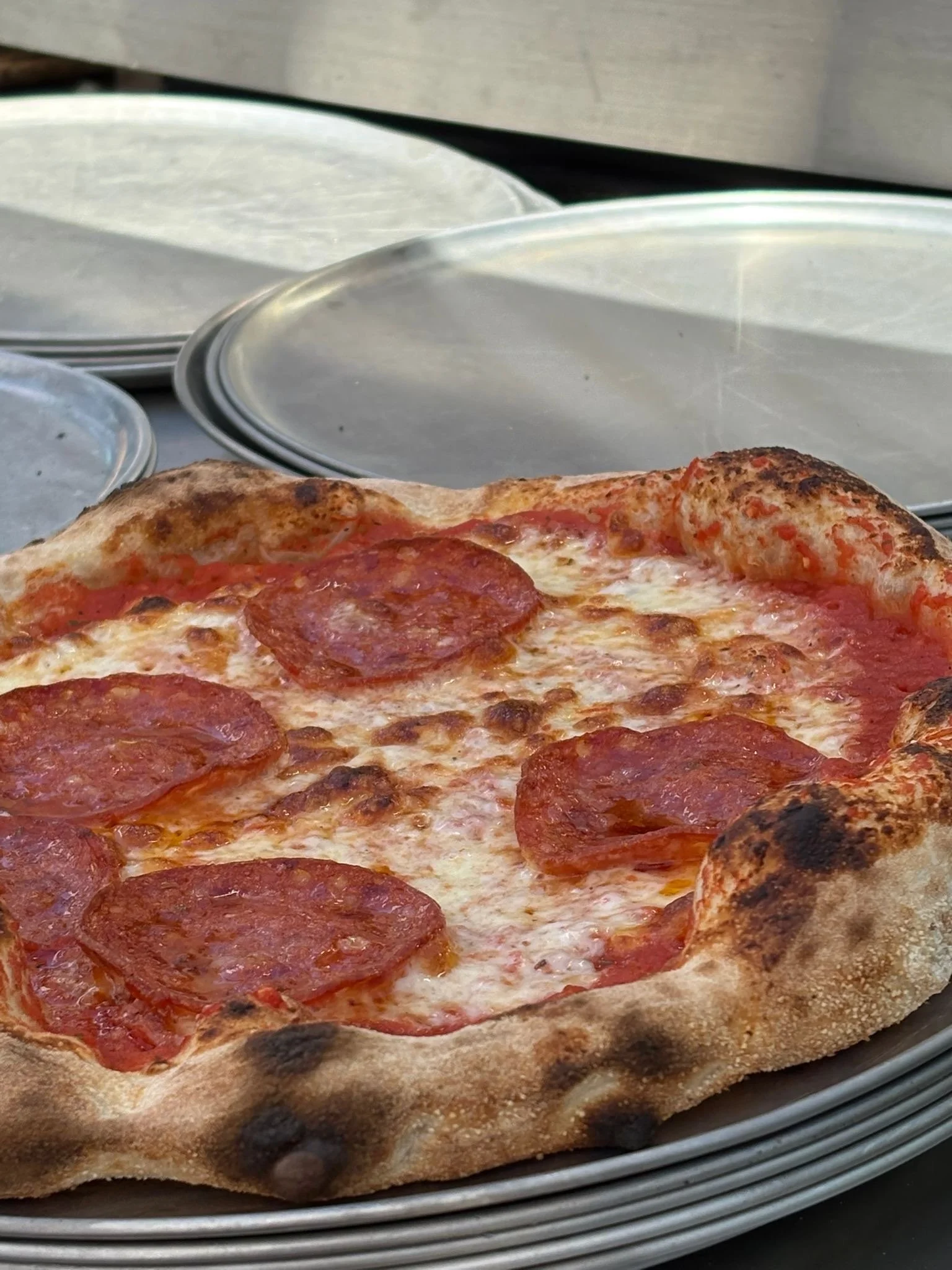 Close-up of a pepperoni pizza with melted cheese and a crispy crust, placed on metal pizza trays with blank metal plates in the background.