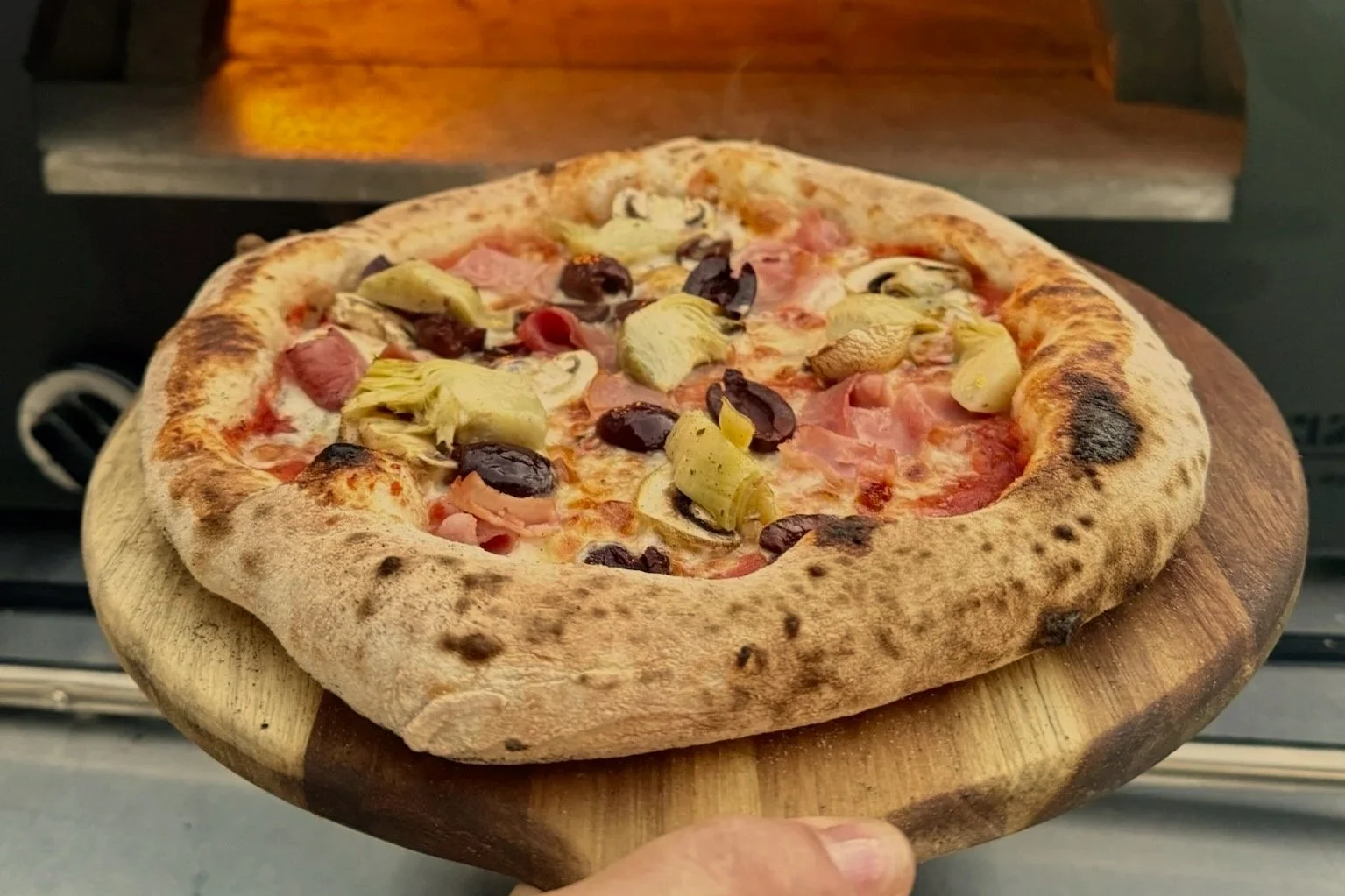 A pizza with black olives, artichoke hearts, and ham on a wooden pizza peel, with a wood-fired oven in the background.