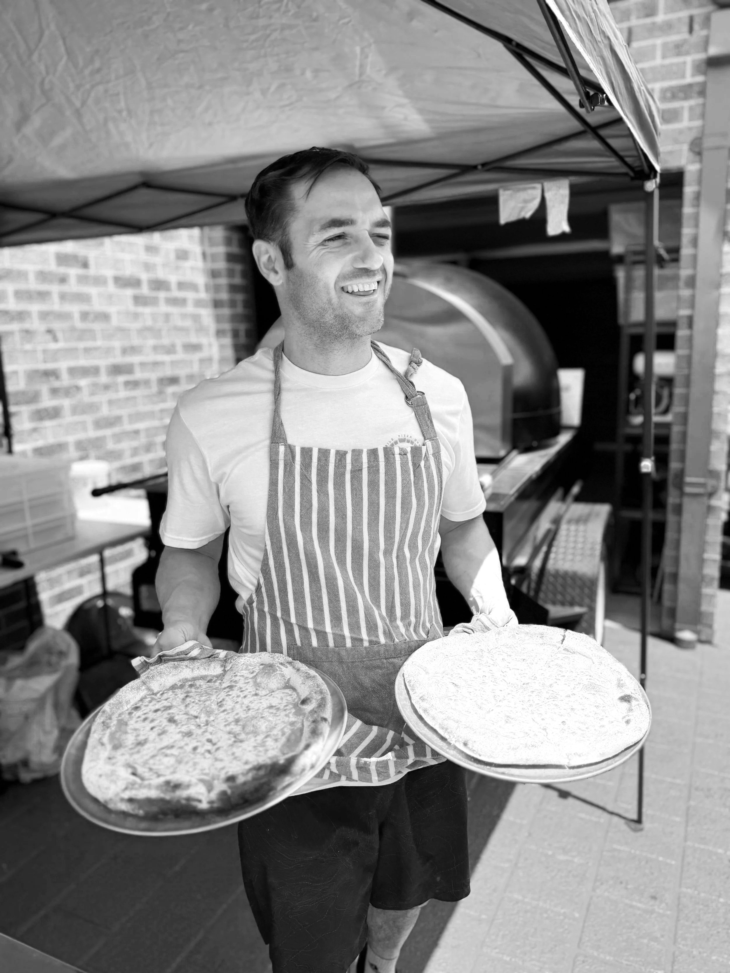 A man smiling and holding two pizzas on trays outside near a brick wall, wearing an apron.