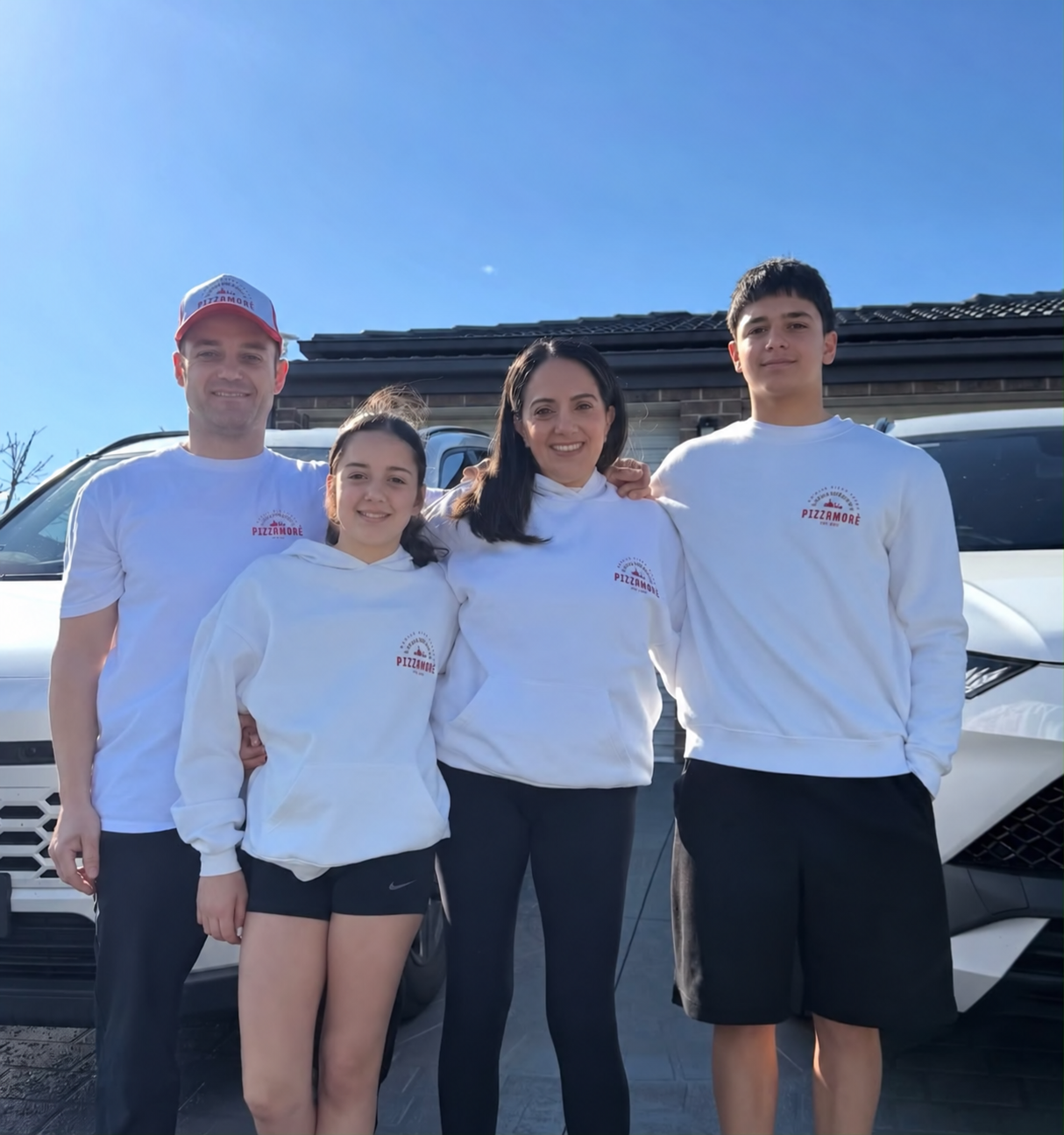 Four people standing outside in front of a white vehicle, all wearing white Pizzamore sweatshirts, smiling at the camera.