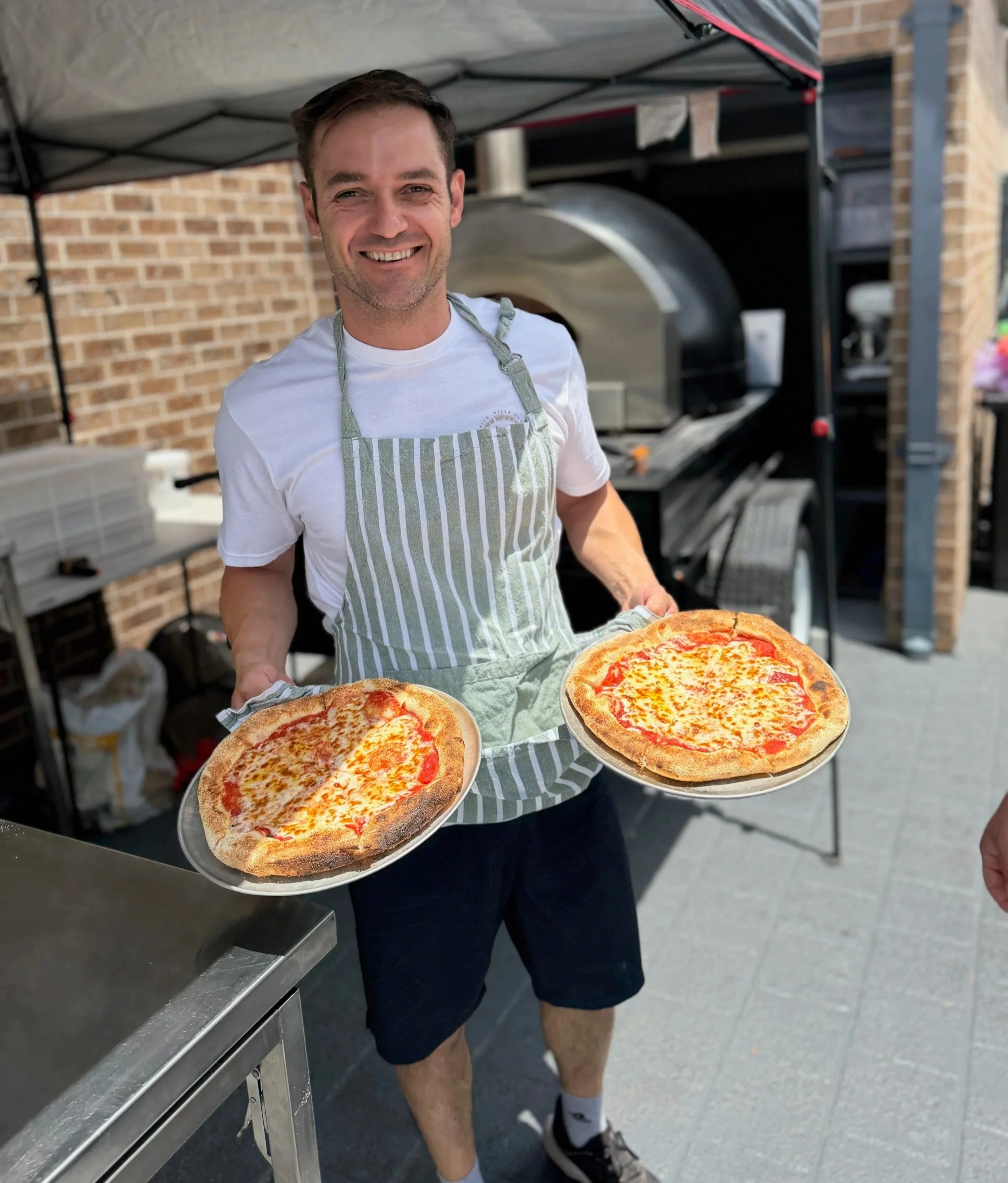 A man wearing a striped apron holding two pizzas on trays, standing in front of a pizza oven outdoors.