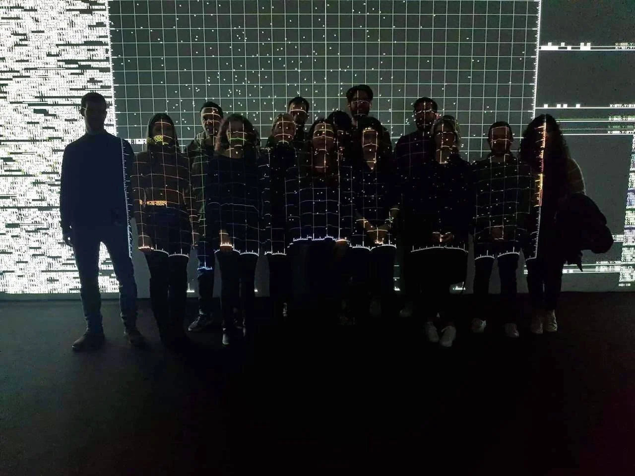 Group of people standing in front of a digital art installation with graphs and data visuals projected on the wall.