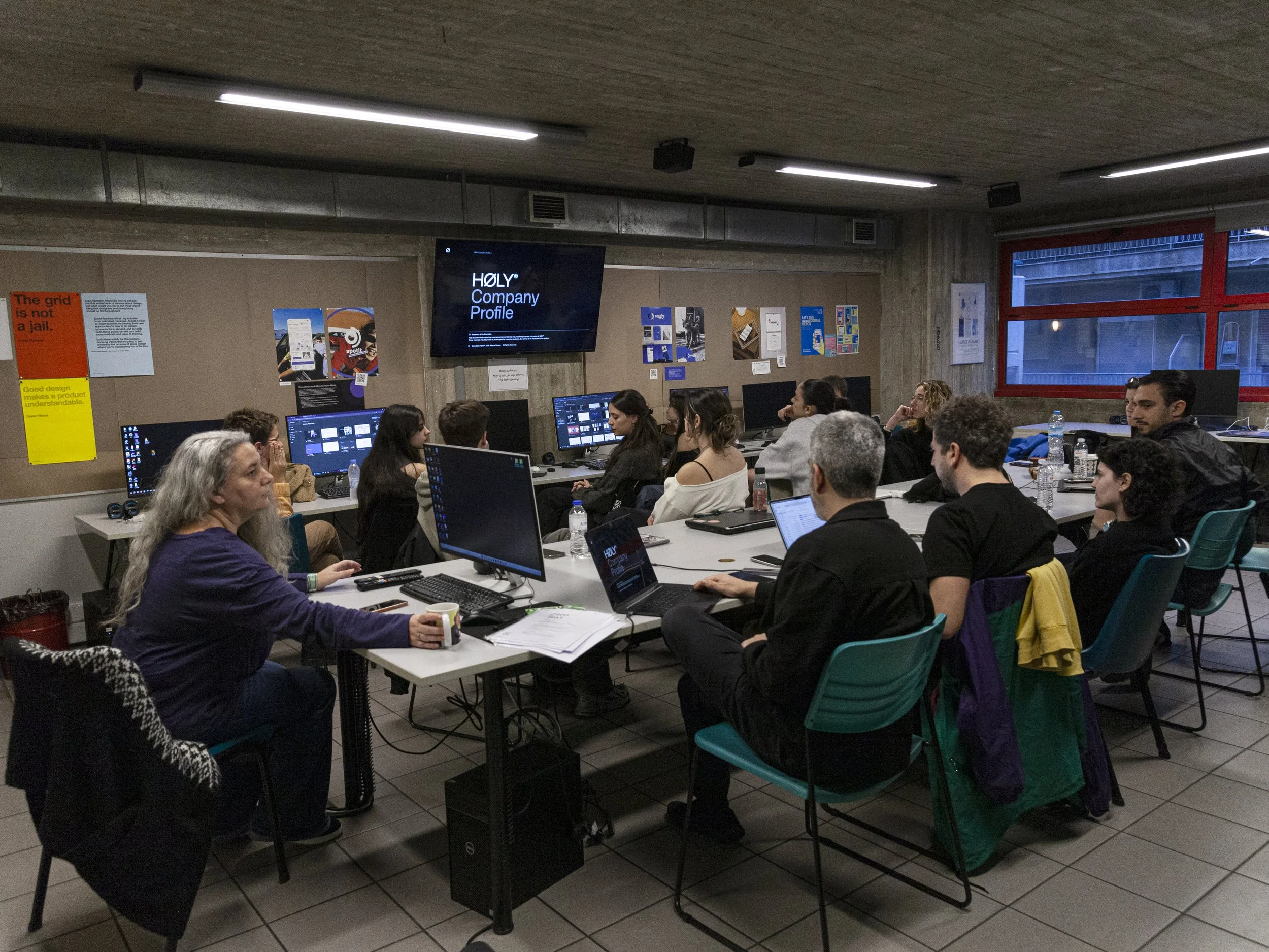 A group of people attending a presentation or class in a room with computers, a large screen displaying 'HOLY Company Profile', and posters on the walls.