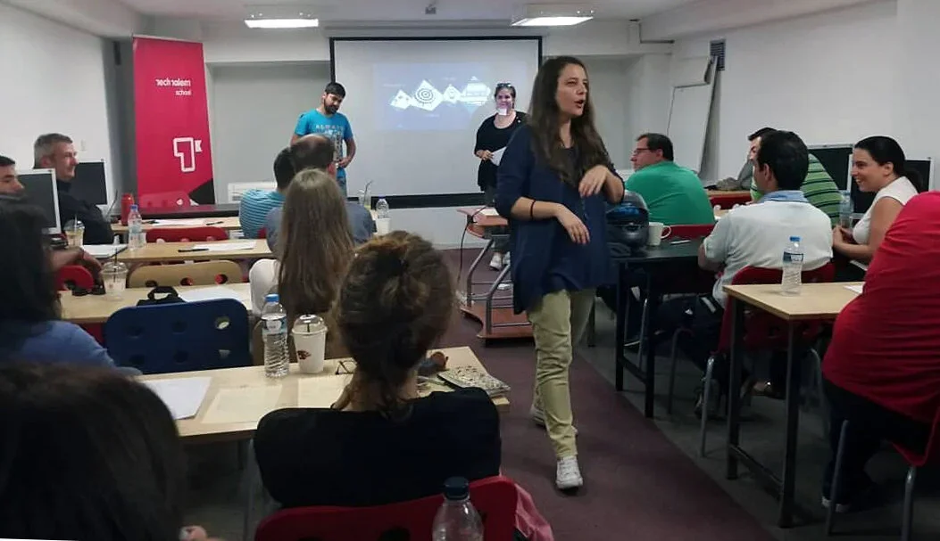 A woman standing and speaking to a group of people seated in a classroom at TechValley School, with a projection screen behind her and people listening attentively.