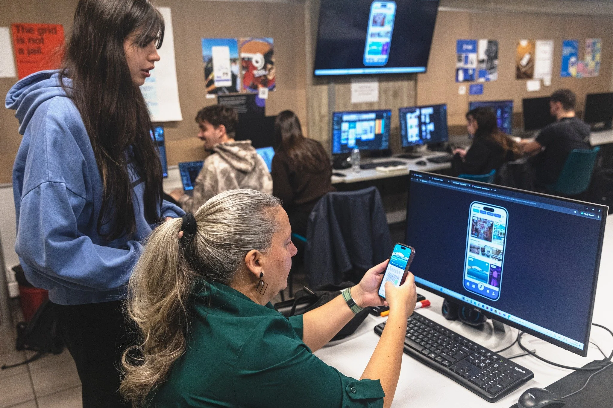 Group of people working on computers in a tech workspace, with a woman showing a phone to a colleague while others work at their monitors.