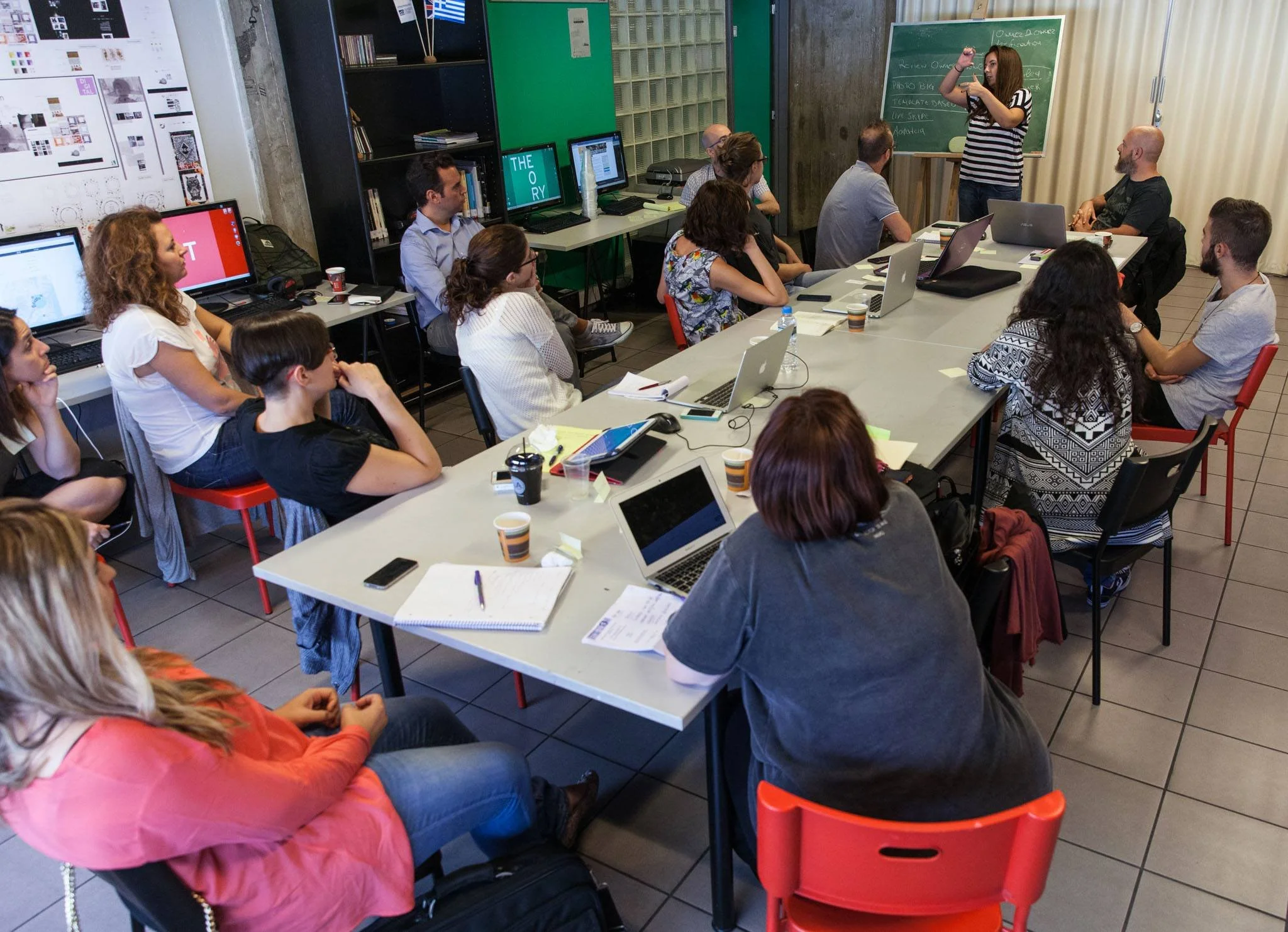A classroom or conference room with a group of people sitting at a long table, watching a woman standing near a blackboard and speaking or presenting. Several laptops and notebooks are on the table. There are also computer monitors, bookshelves, and posters on the walls.