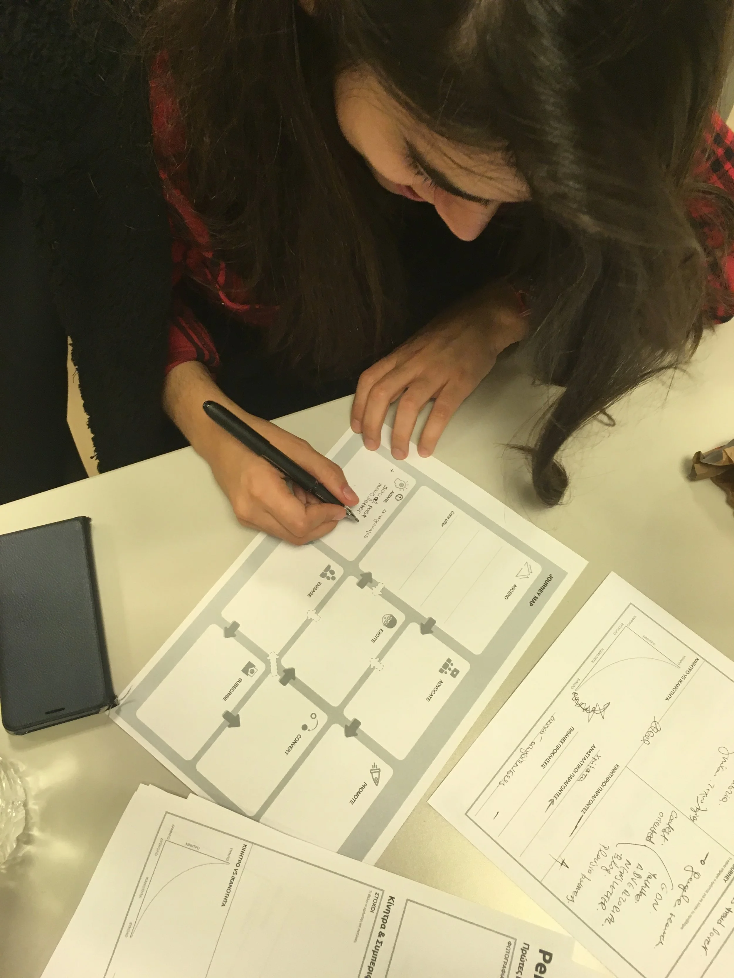 A woman with long dark hair writing on a board game or diagram at a table, with papers and a black phone nearby.