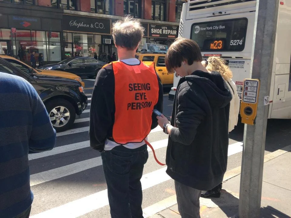 Two young people standing on a city sidewalk at a crosswalk, one wearing an orange vest with the words 'Seeing Eye Person' and looking at his phone, the other a girl with glasses and a hoodie, also looking at her phone. There are cars and a city bus in the background.