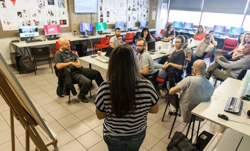 A woman presenting to a group in a classroom or office setting with participants sitting at desks, computers, and windows with blinds in the background.