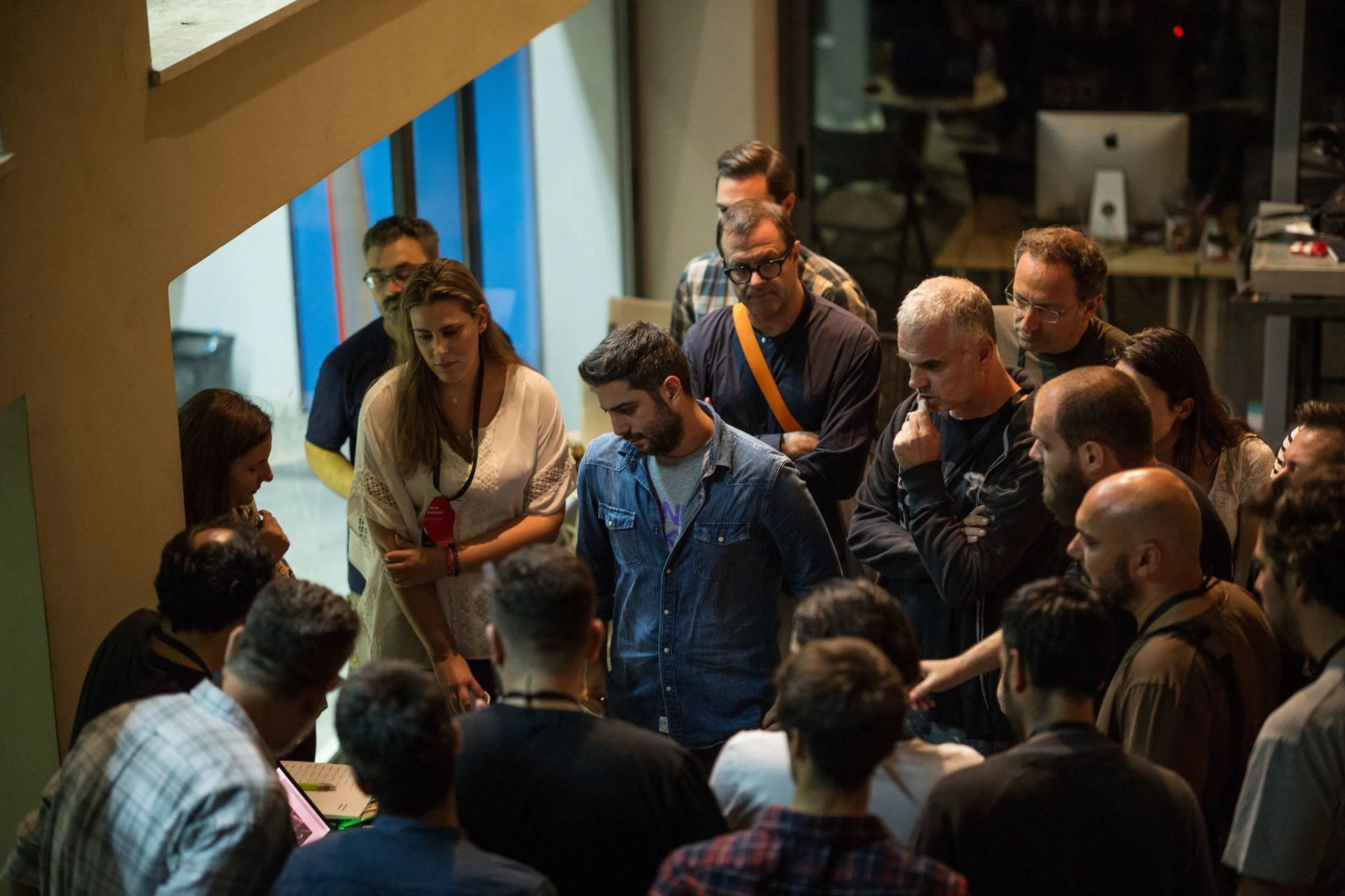 Group of people gathered around a table at an indoor event, looking at a laptop or display, with some standing and some sitting, in front of a large window and behind a modern office space.