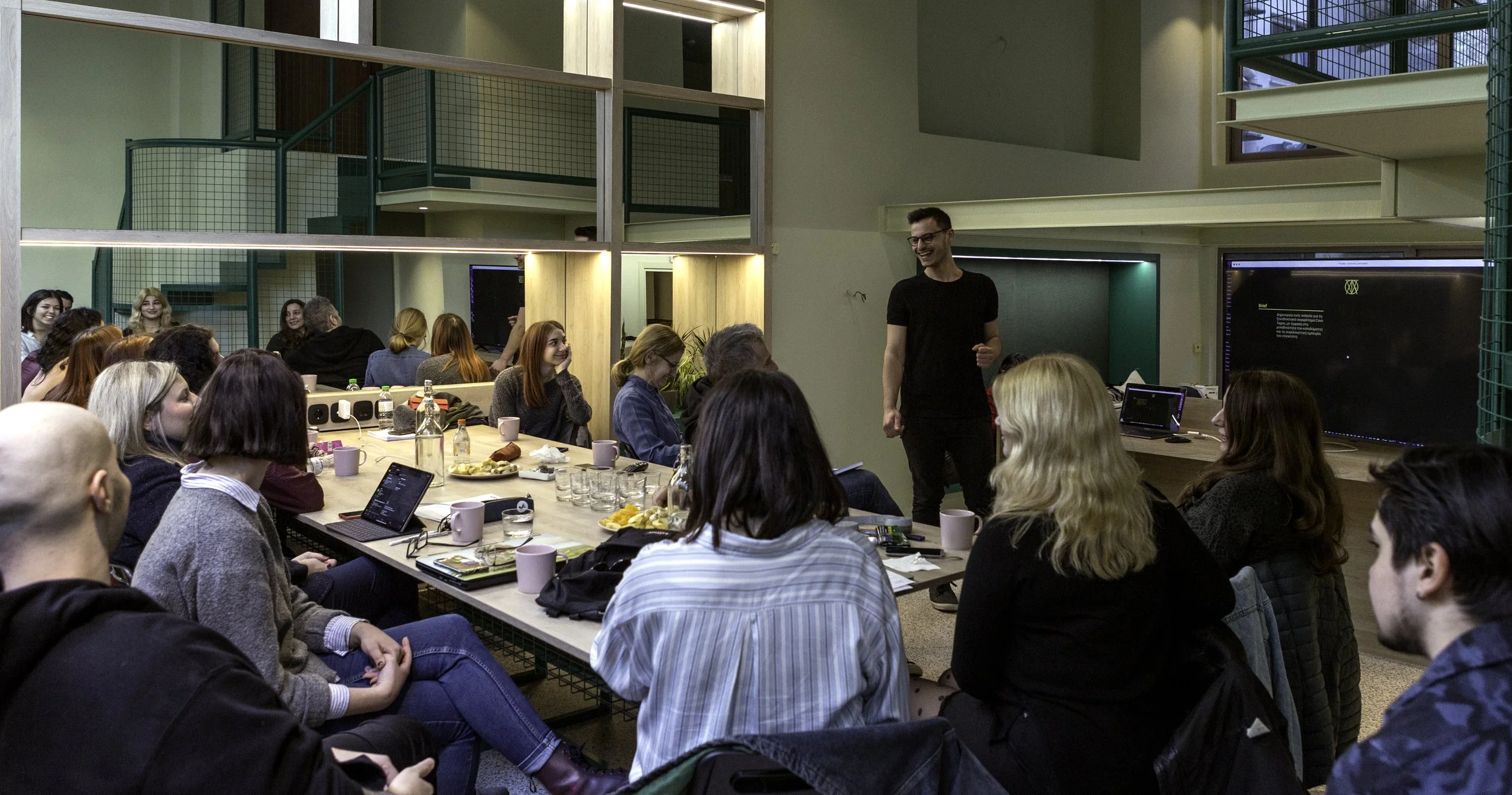 A man standing and smiling at a group of people seated at a long table in a modern conference room. The table has laptops, food, drinks, and notebooks. The room has a two-story structure with stairs, large windows, and a large screen on the wall.