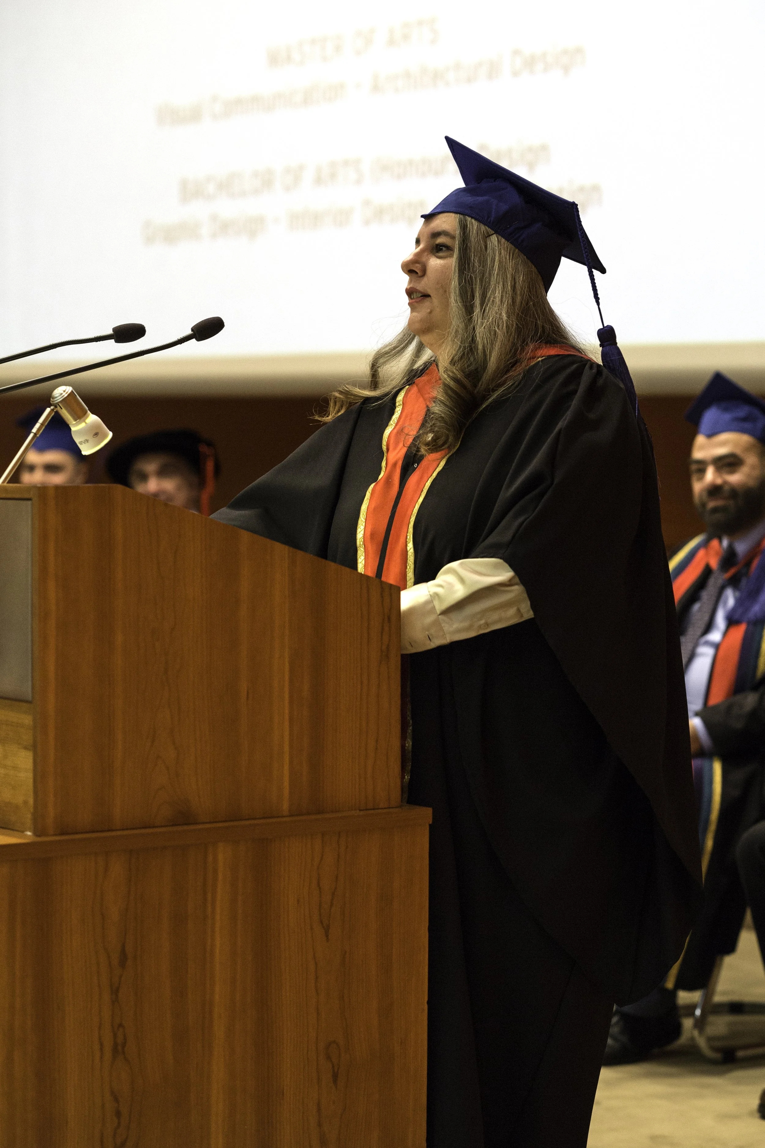 A woman in a black graduation gown and blue cap speaking at a podium during a graduation ceremony, with other graduates seated in the background.