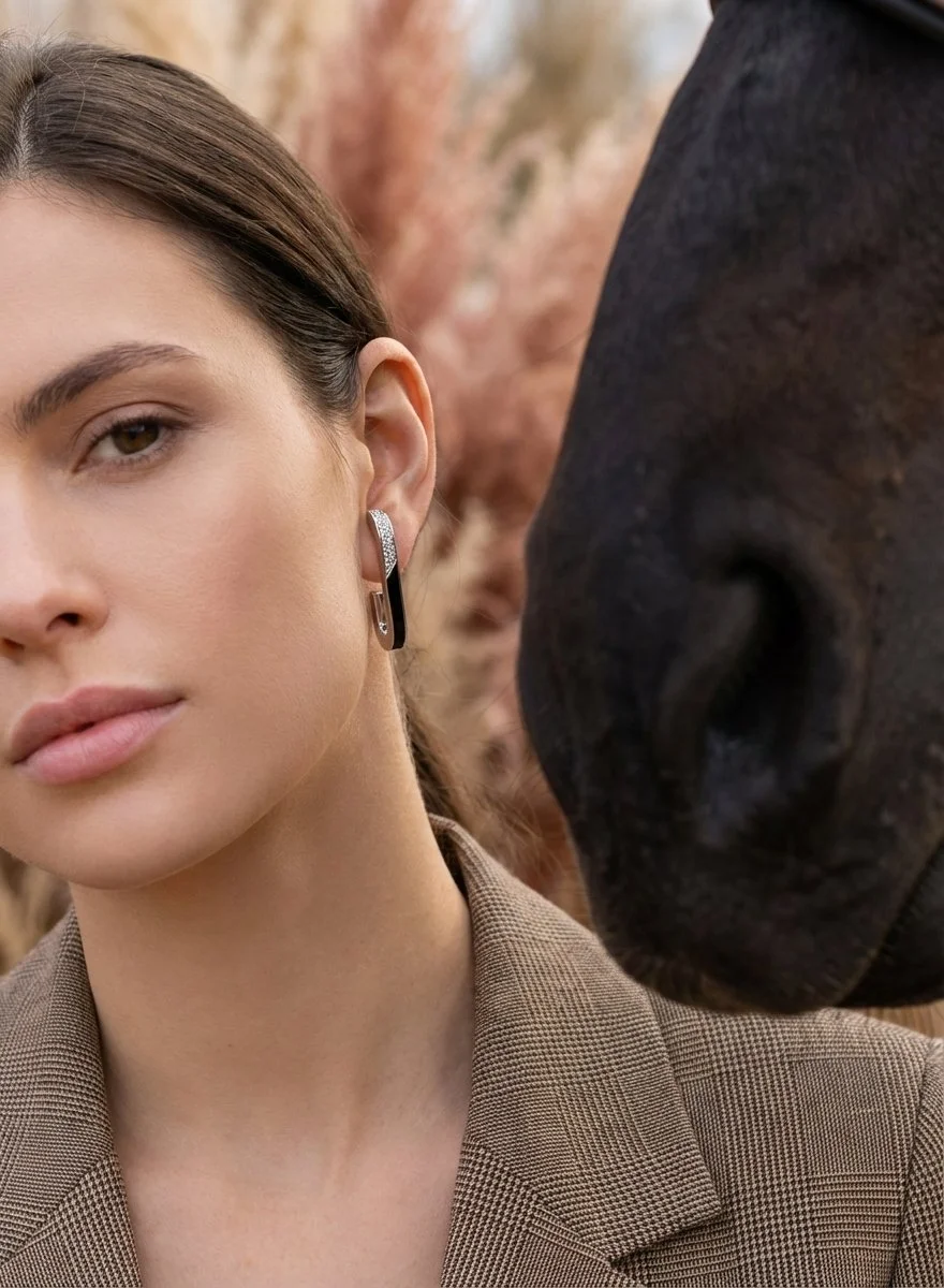 Close-up of a woman with brown hair wearing a light brown blazer and half onyx half diamond earrings with the top half looped into her ear like a cuff and the other half hanging down, standing next to a horse in the meadows.