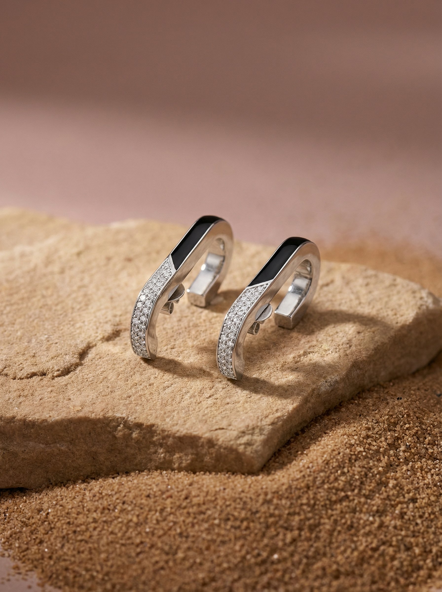 Pair of white gold earrings with onyx and diamond embellishments on a sandy beige surface with soft pink background.
