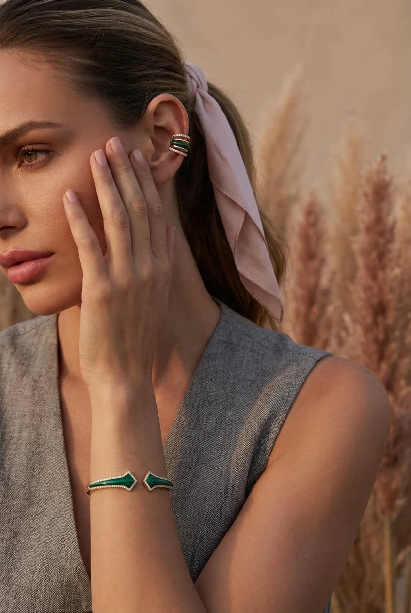 A portrait of a woman with light brown hair wearing a light pink bow in her hair tied up, a grey sleeveless top, and matching sterling silver in yellow gold vermeil and malachite 5 ring ear cuff with arrowed bracelet pointing to each other. 