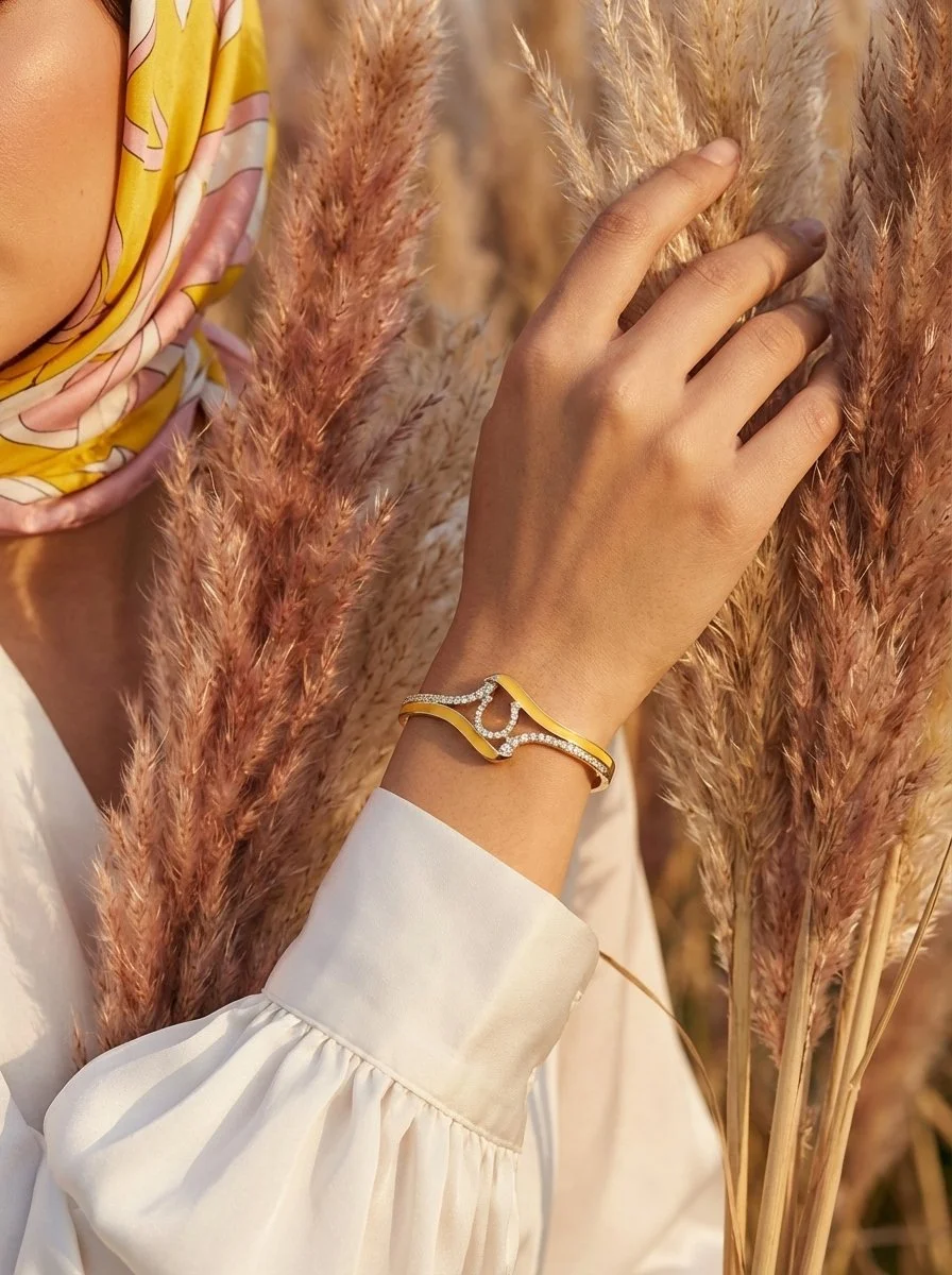 A woman's close up hand shot with a horseshoe in the middle of a yellow and gold bracelet resting on tall, pinkish pampas grass. The person is wearing a white long-sleeve shirt with gathered cuffs and a patterned headscarf in yellow and pink.