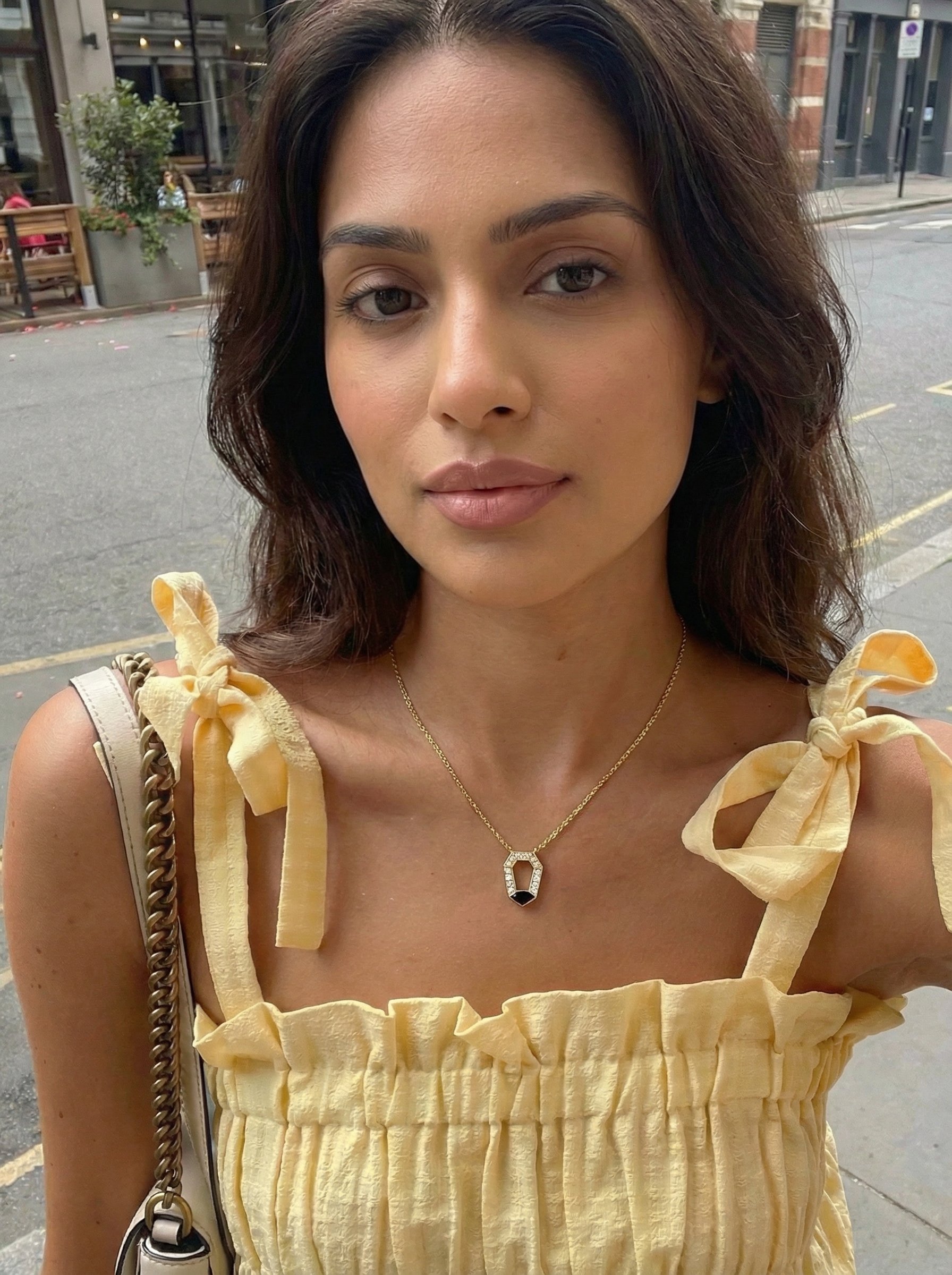 A young woman with light brown skin and dark wavy hair is standing outdoors on a city street, wearing a yellow top with tied shoulder straps and a gold necklace with a black pendant.