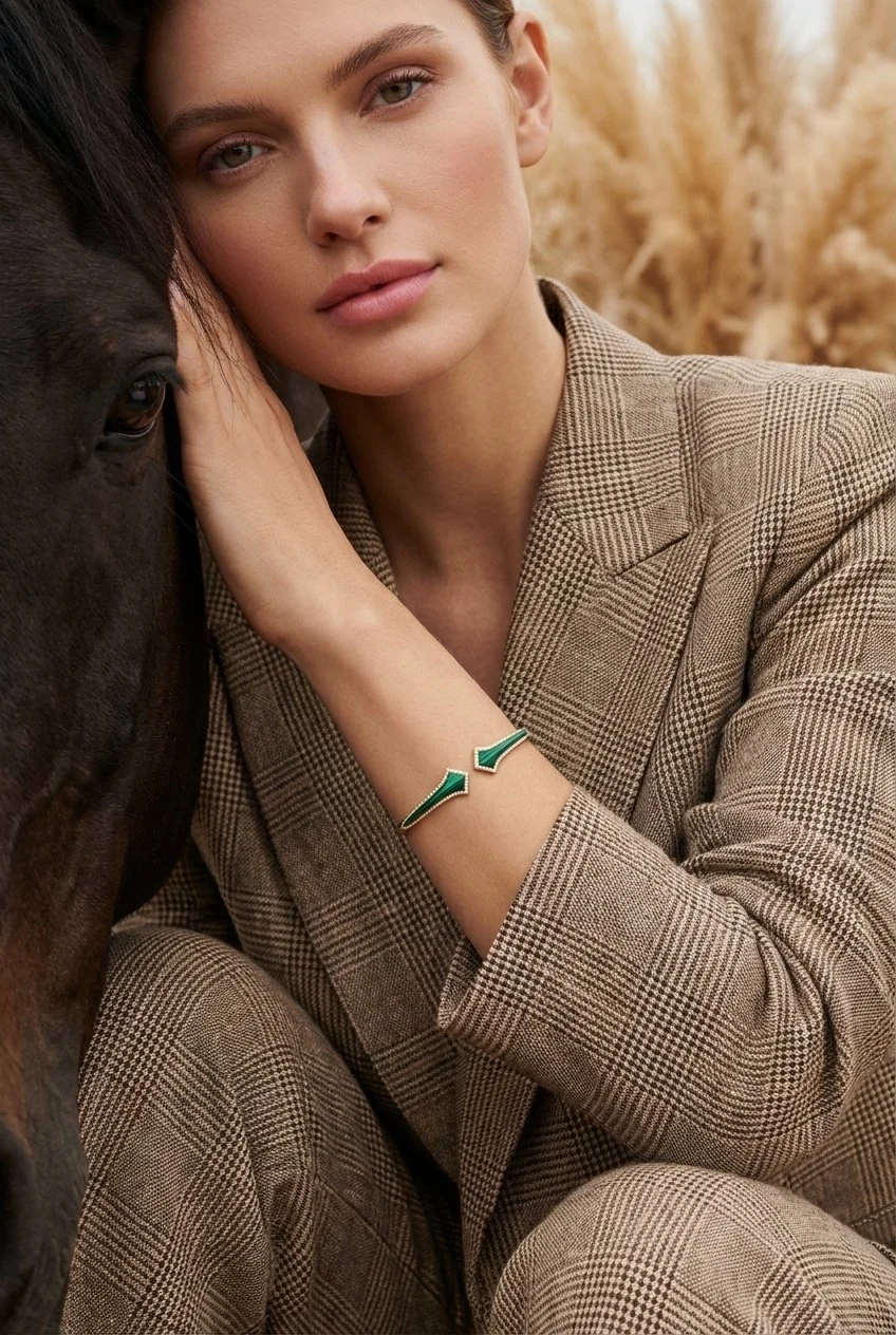 A portrait shot of a woman with fair skin and blonde hair, dressed in a brown plaid suit, rests her head against a black horse in the meadows, wearing a sterling silver bracelet in yellow gold vermeil of two arrows pointing each other in malachite.