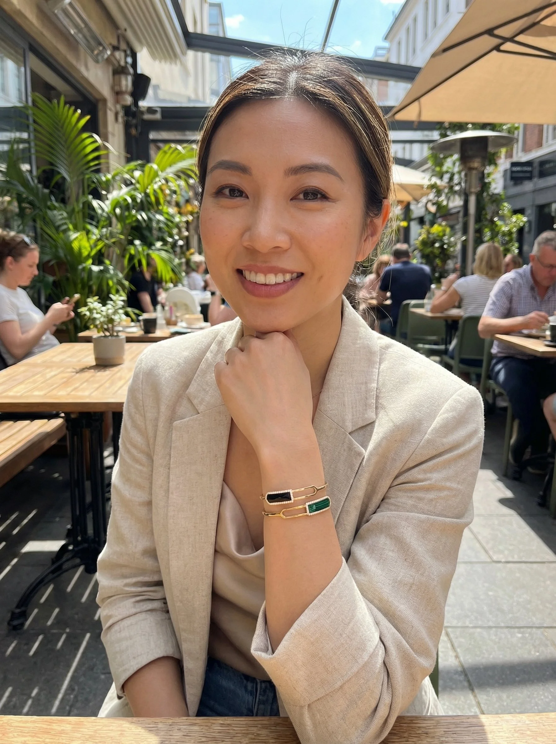 A woman sitting at an outdoor cafe, smiling at the camera. She has a bangle that is designed to give the illusion of 2 bangles but is 1. The piece has two rectangular stones with one pointy end facing outdoors, one in onyx and another in malachite.
