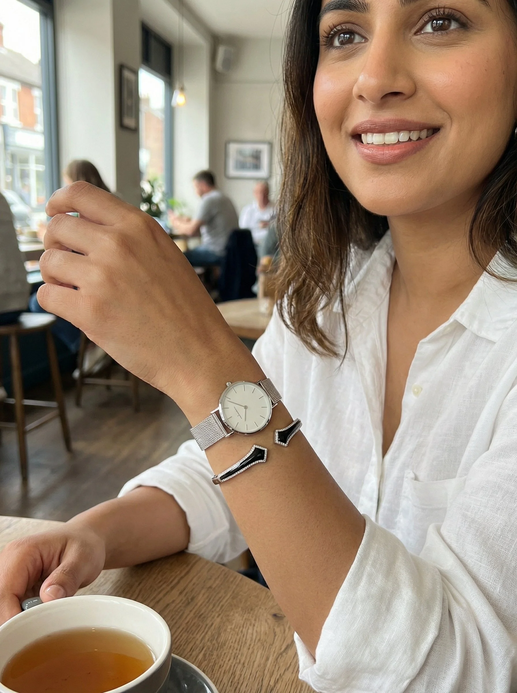 A woman sitting at a cafe table with a cup of tea, wearing a white shirt, watch, and a full onyx bangle of arrows facing each other, smiling near large windows with other people in the background.