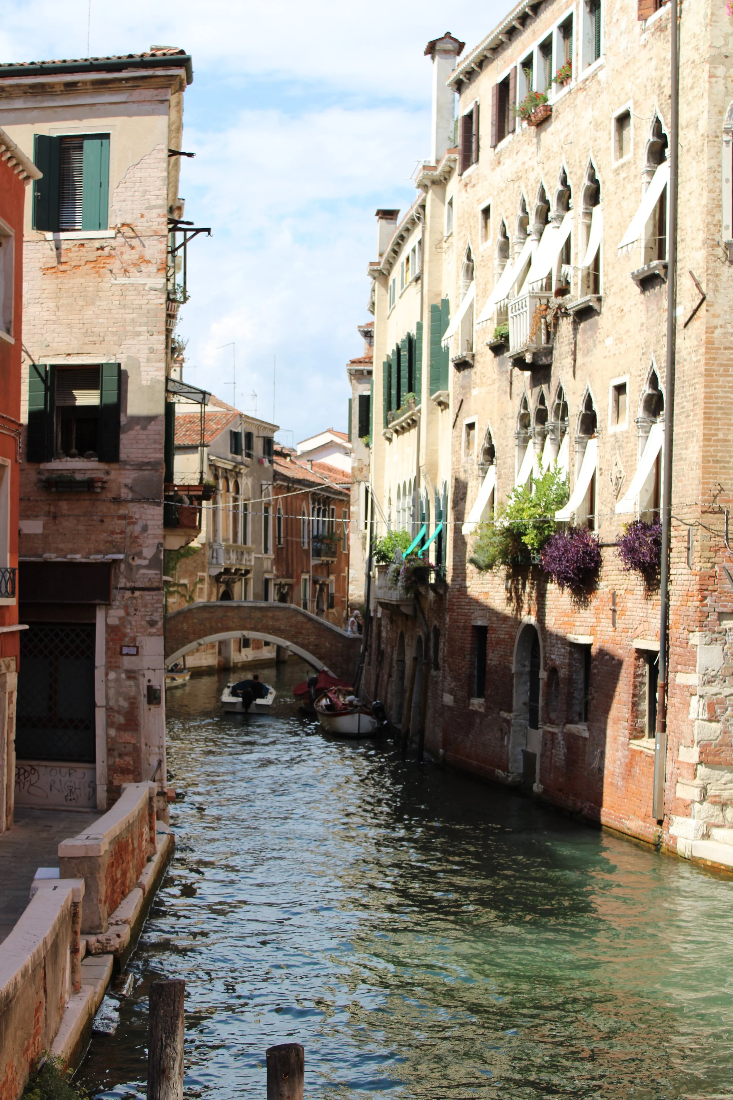 A canal scene in Venice, Italy, with old buildings on either side, a small stone bridge in the background, and boats floating on the water.
