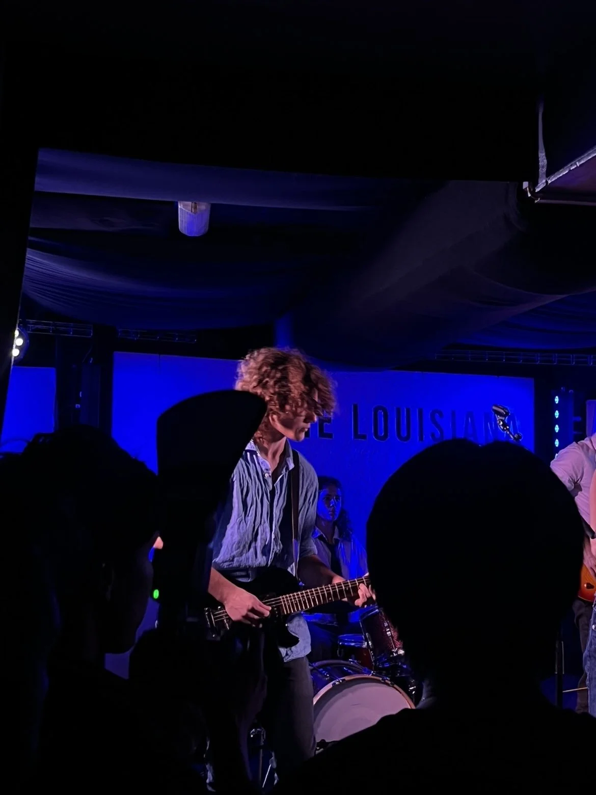 A musician playing an electric guitar on stage during a live performance, with a large drum in the background and audience silhouettes in the foreground.