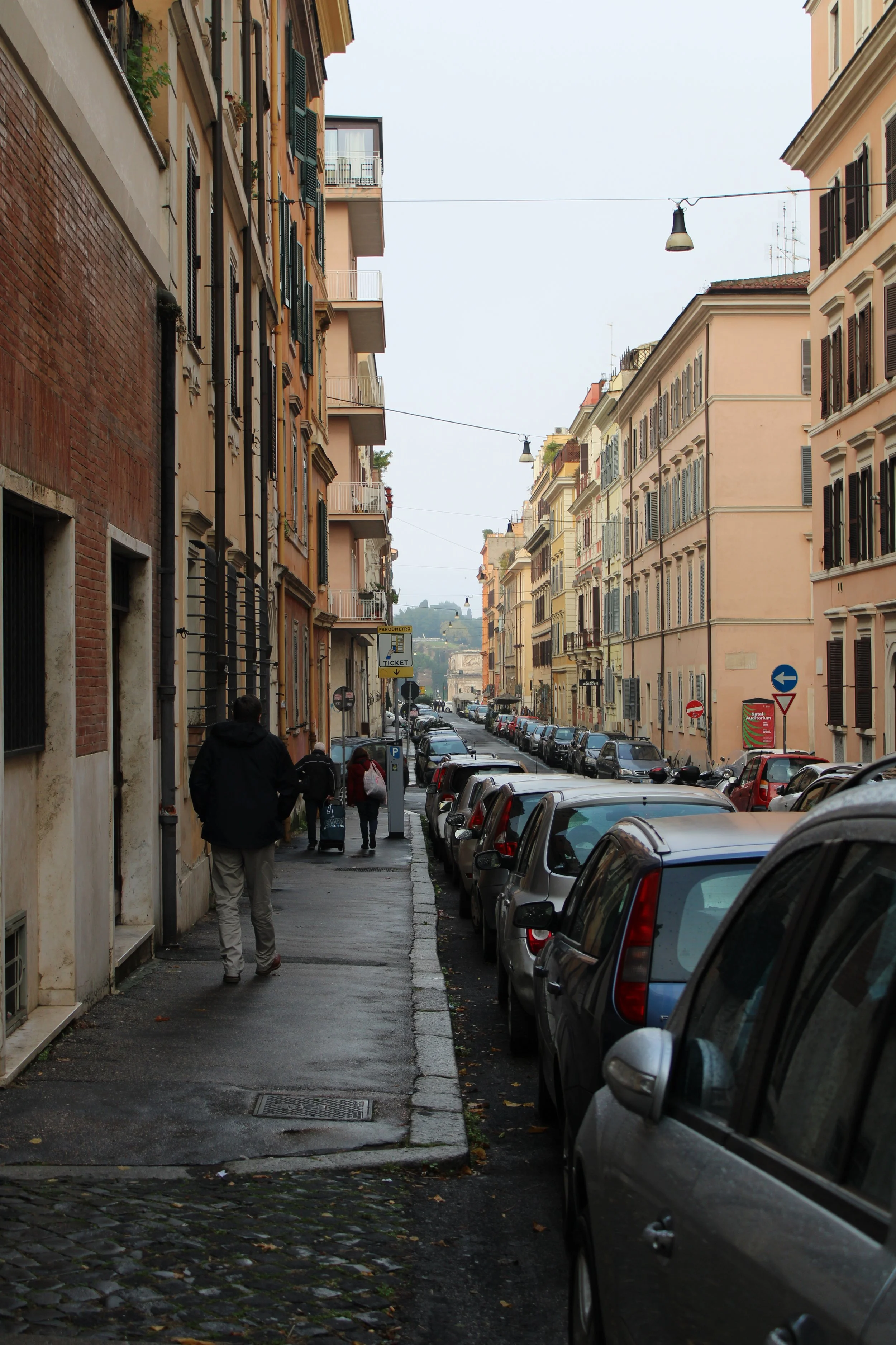 A narrow street scene in Europe with parked cars lining the sidewalk and pedestrians walking through. Colorful old buildings with green shutters tower above.
