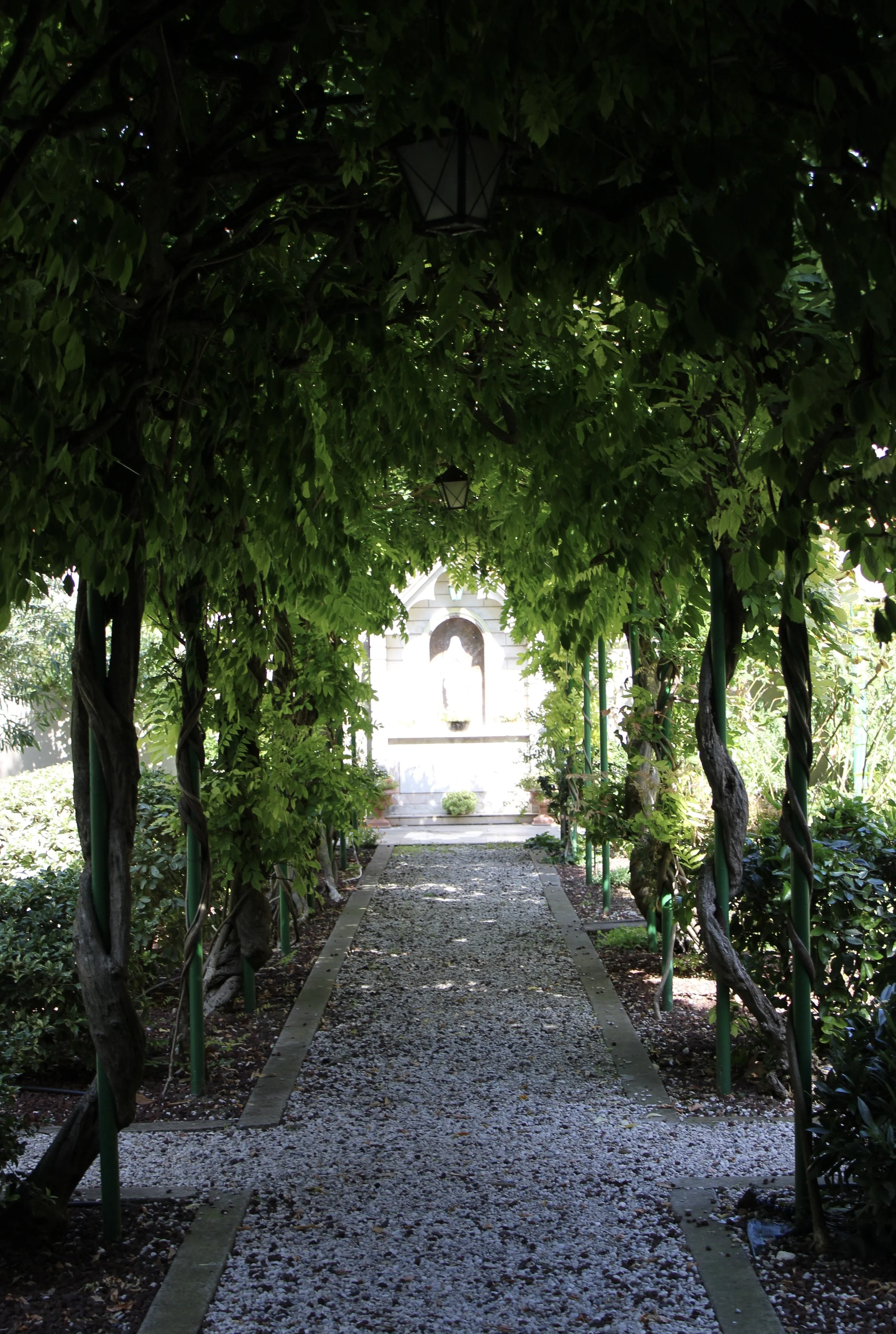 A shaded garden walkway with a gravel path, green leafy arch overhead, leading to a white religious statue or monument at the end.