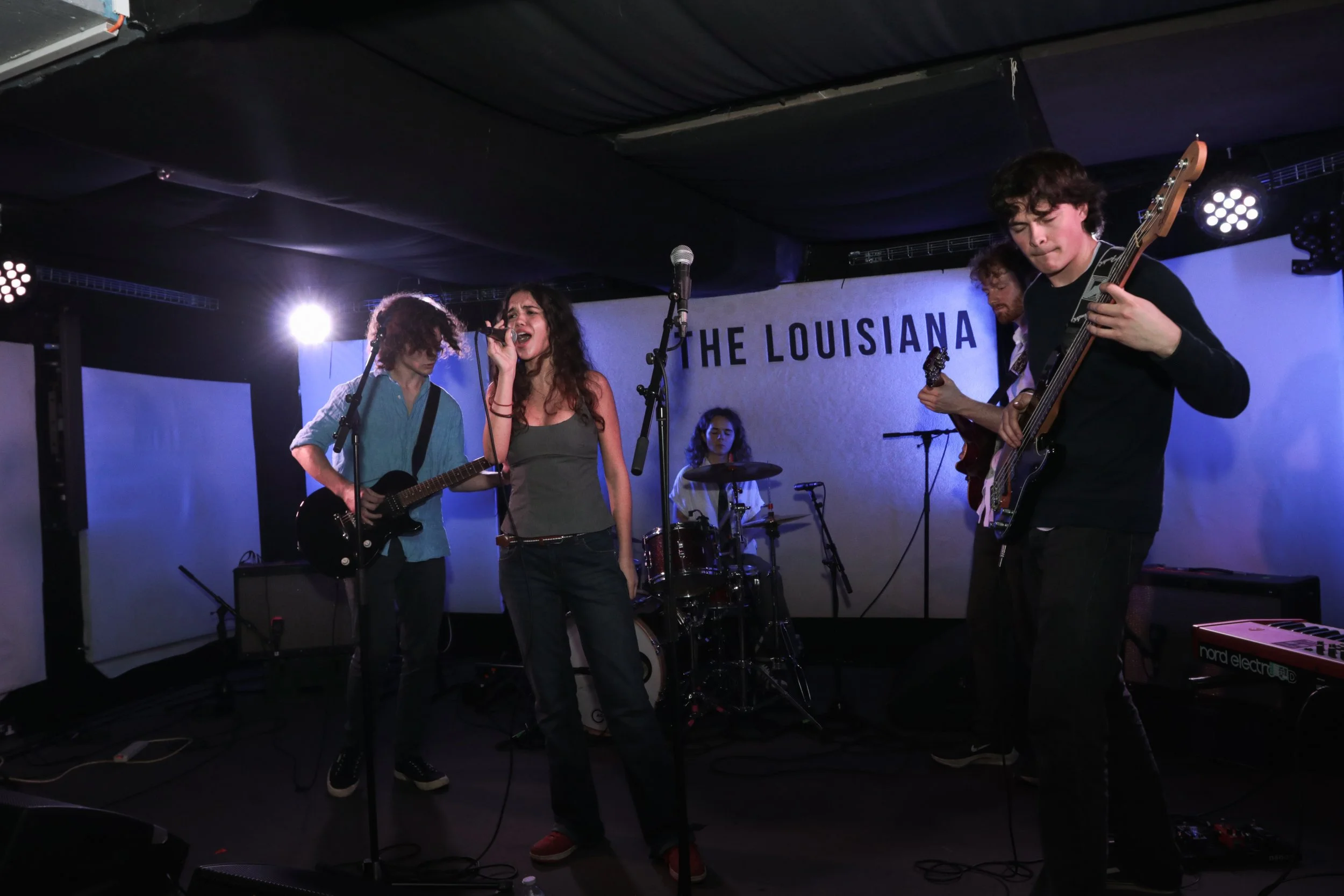 A band performing on stage with four musicians, including a female singer and a drummer, in a dimly lit venue. The backdrop reads "THE LOUISIANA."