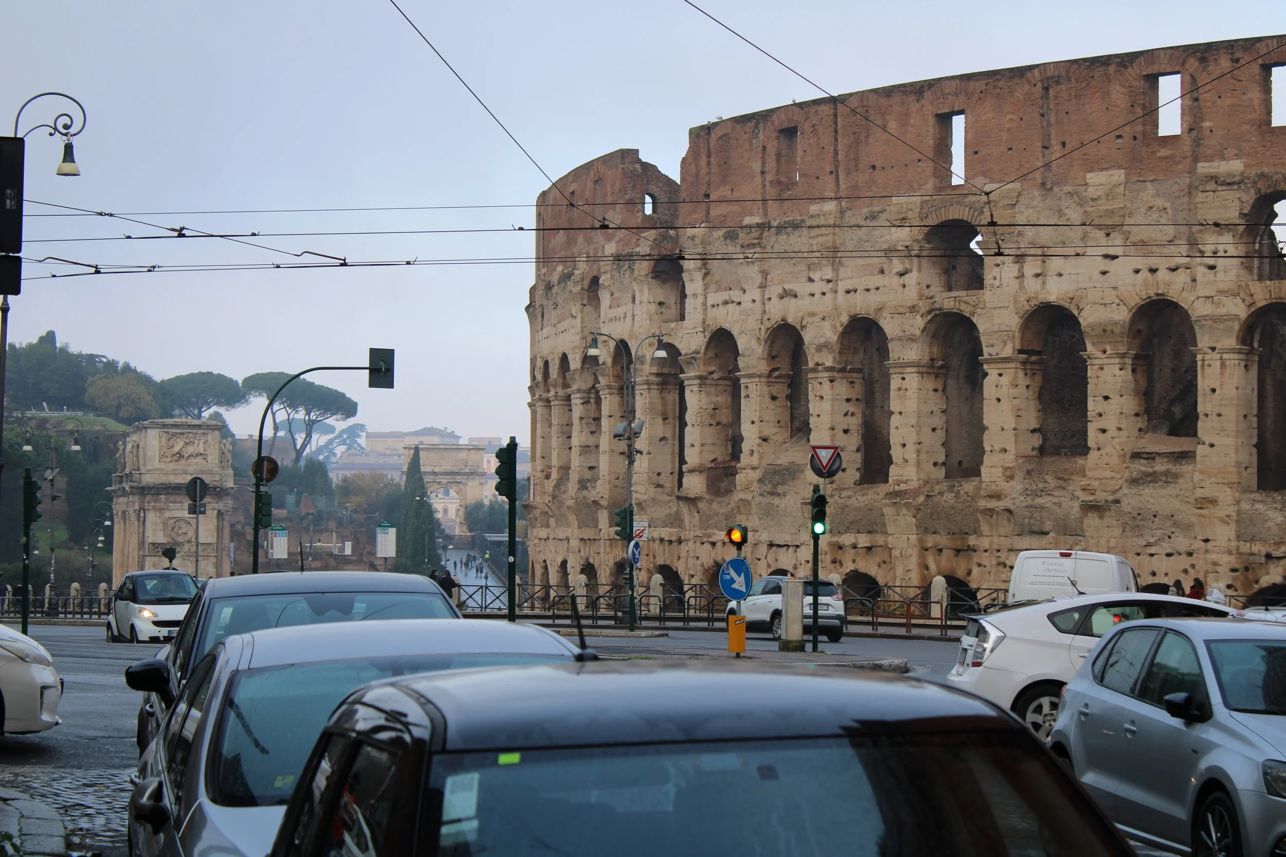 The ancient Roman Colosseum in Rome, Italy, with cars and traffic lights in the foreground and trees in the background.