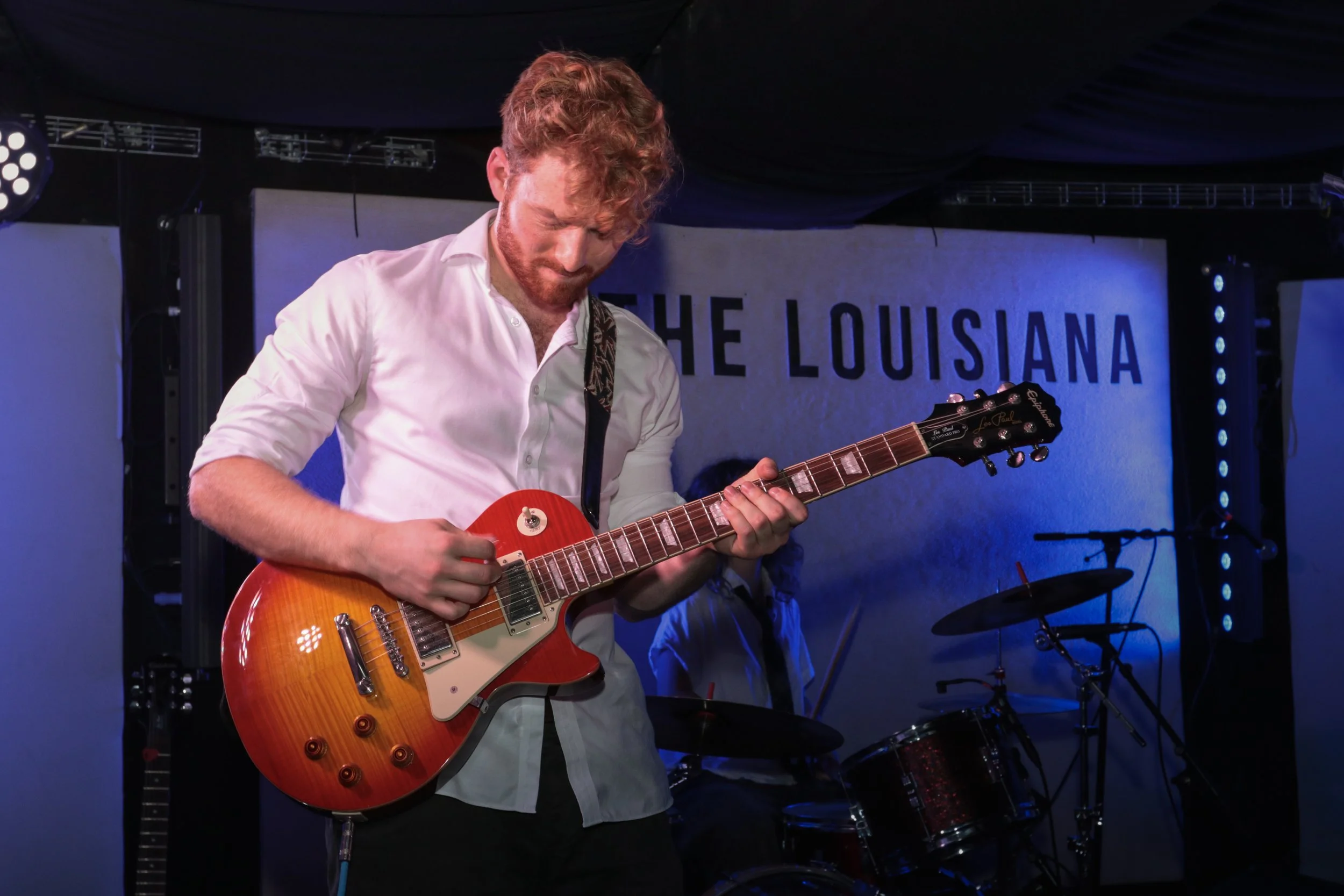 A man with curly red hair and a beard playing an orange electric guitar on stage with a dark background and a sign that reads "The Louisiana".