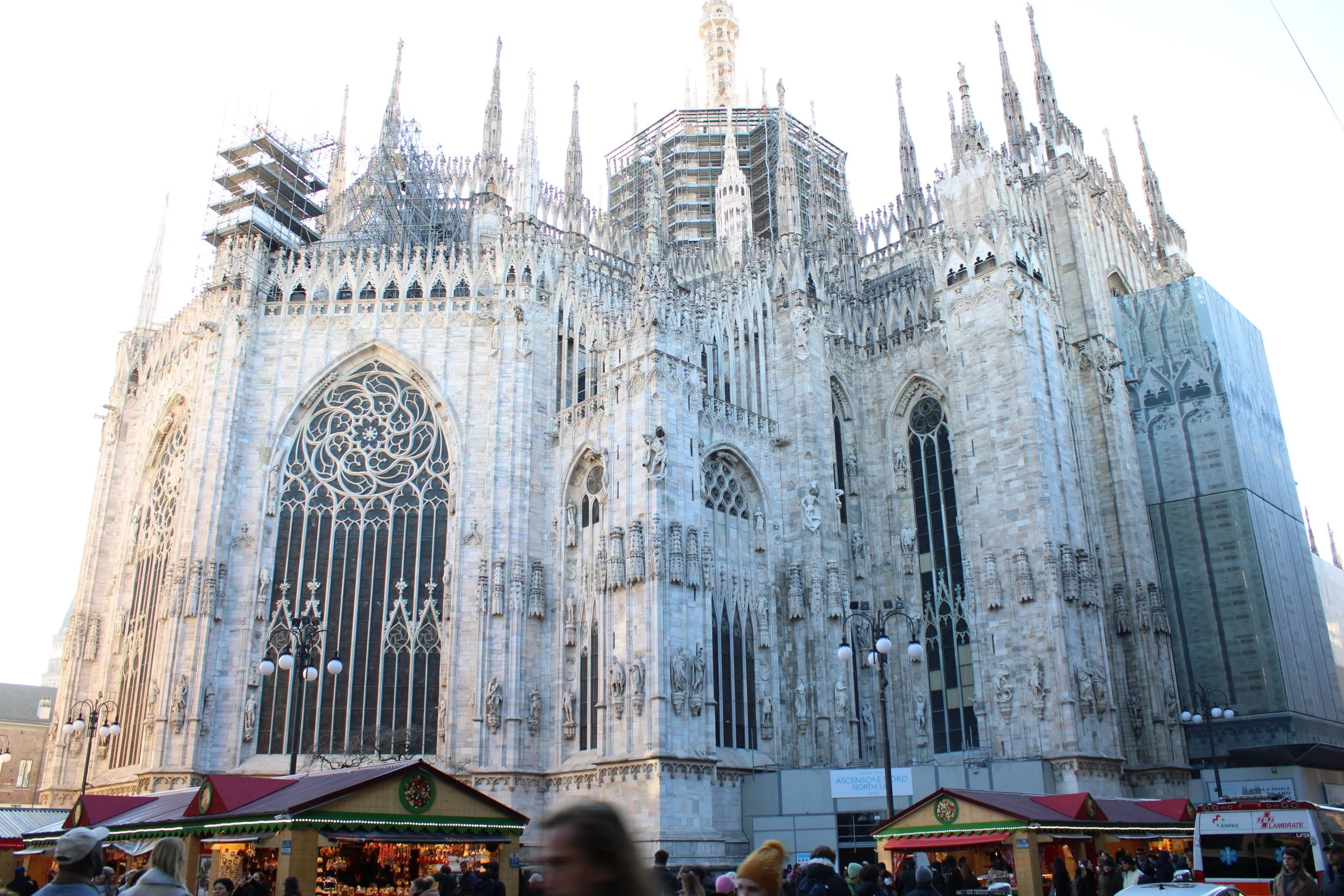 View of the Milan Cathedral (Duomo di Milano) with Christmas market stalls in the foreground and people walking by.