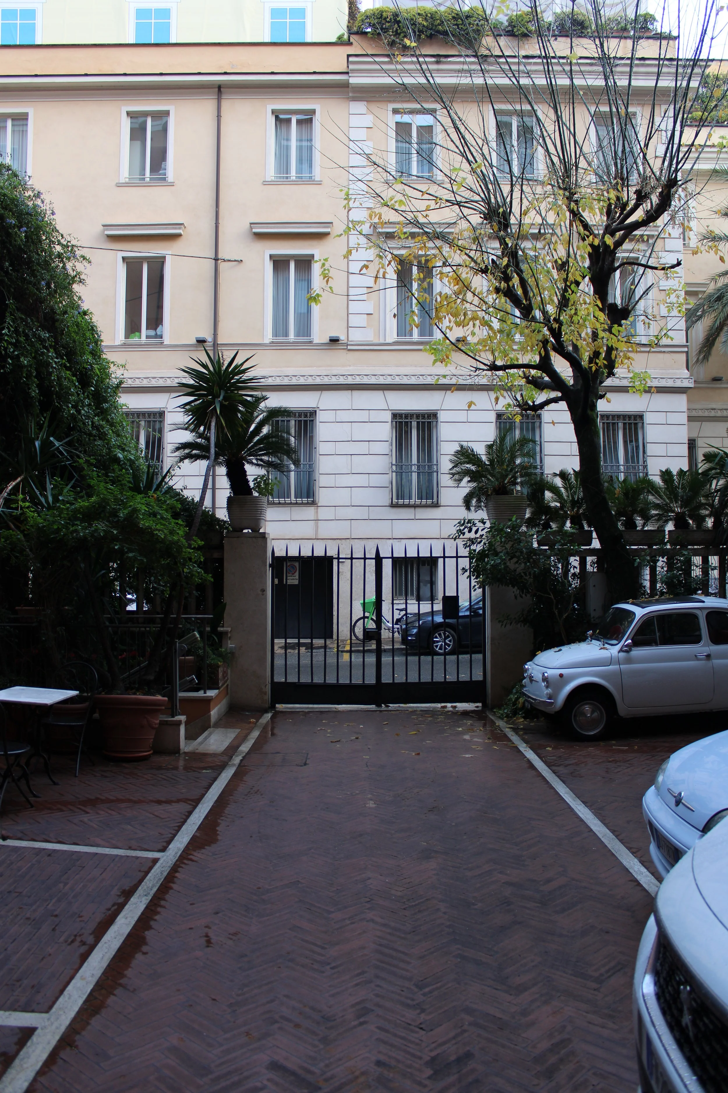 Urban courtyard with parked cars, trees, potted plants, and a gated entrance, with apartment building in the background.