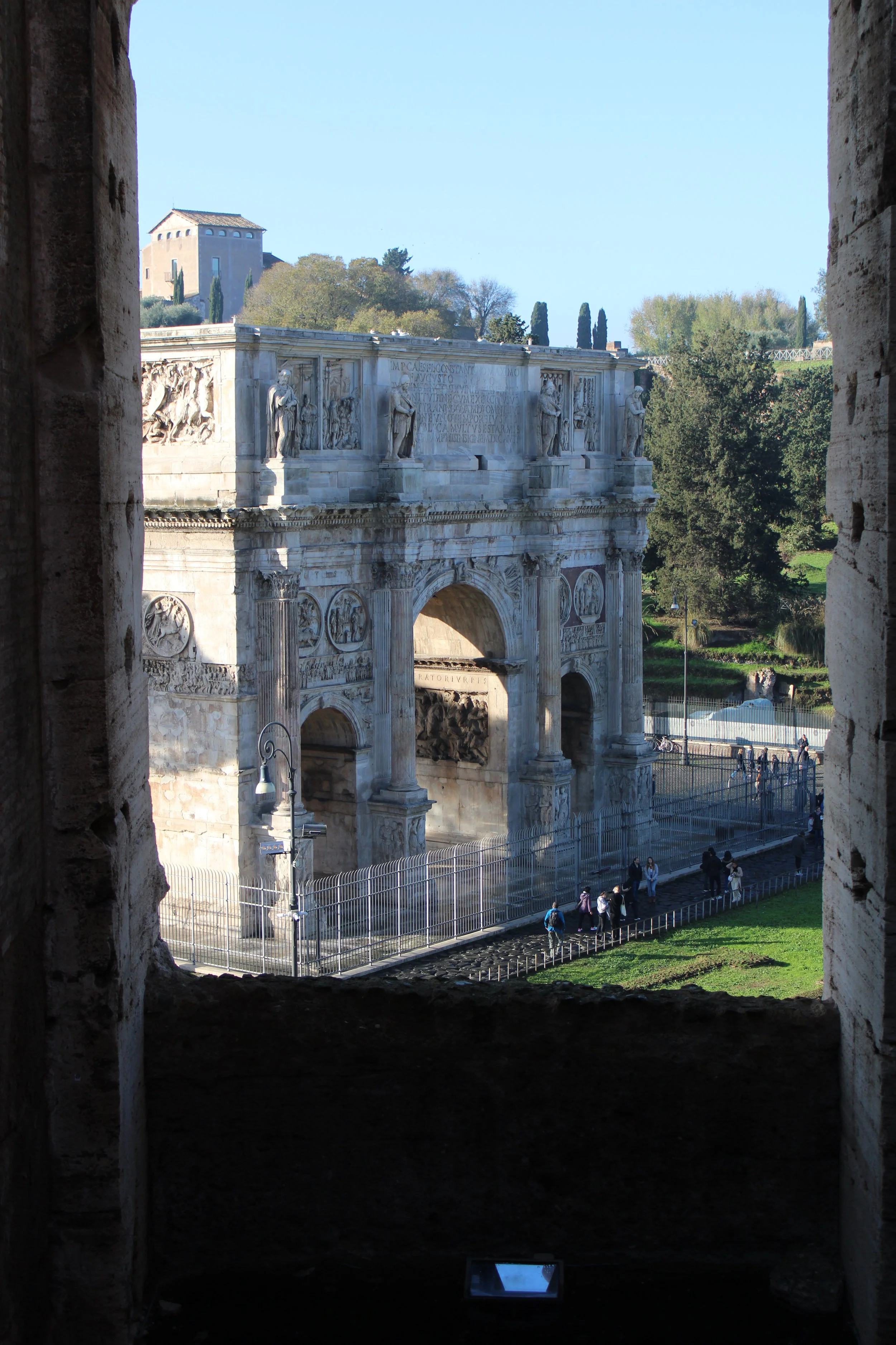 View of the Arch of Constantine in Rome, Italy, through a window, with tourists walking nearby and trees in the background.