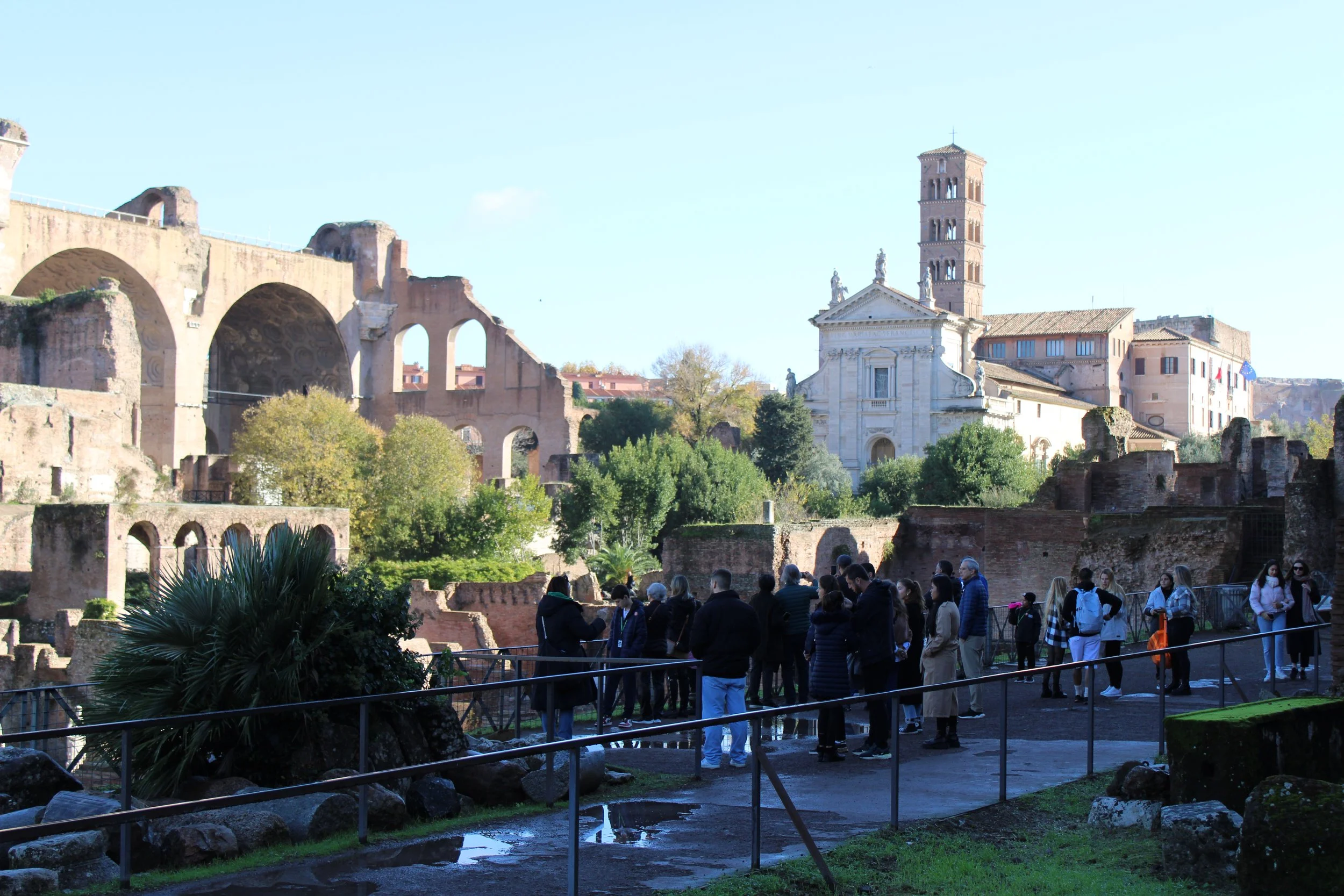 Tourists visiting the ancient ruins of the Roman Forum in Rome, Italy, with historic arches and a church with a bell tower in the background on a clear day.