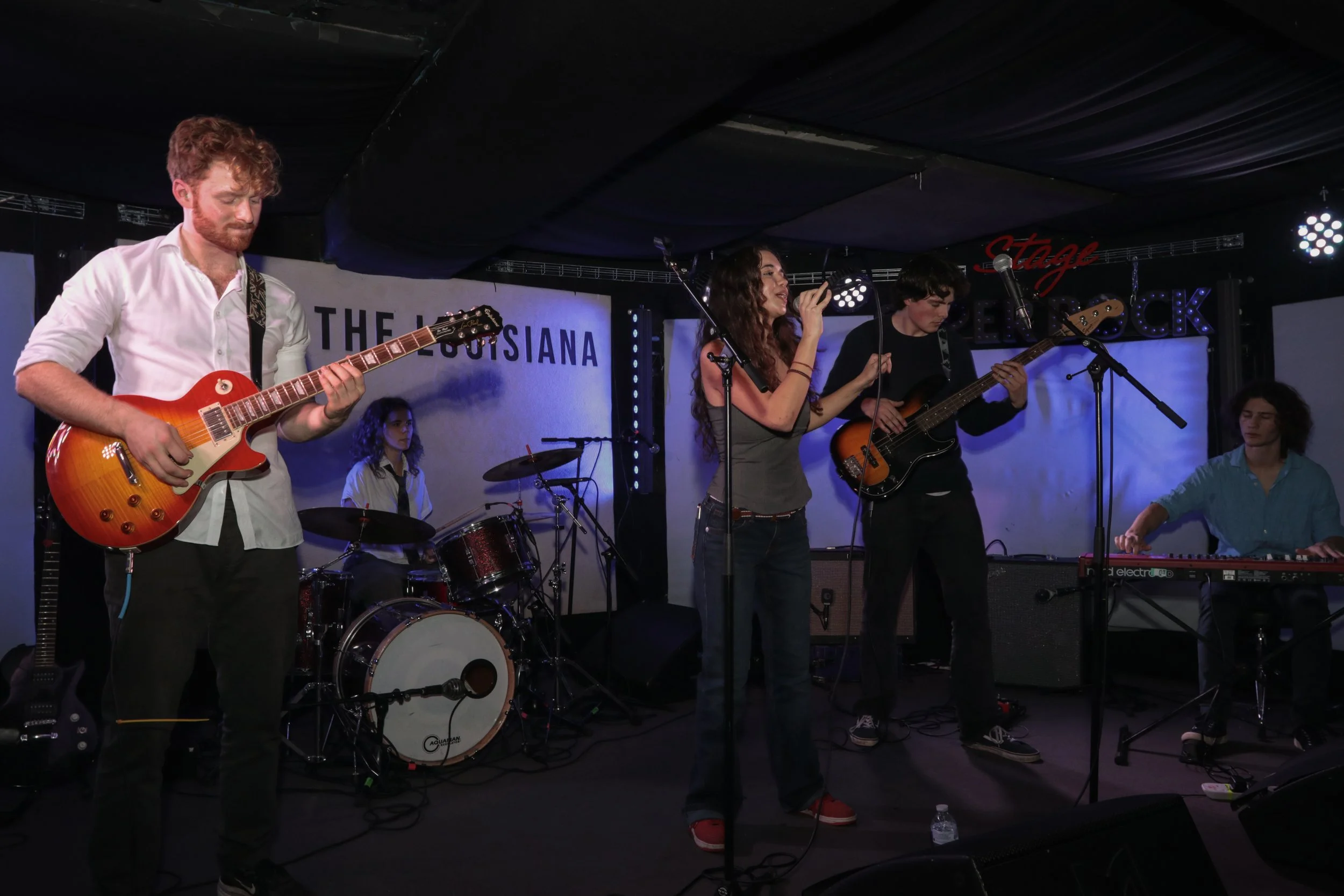 A band performing on stage, with five members: a guitarist, a drummer, a vocalist, a bassist, and a keyboardist, in a music venue.