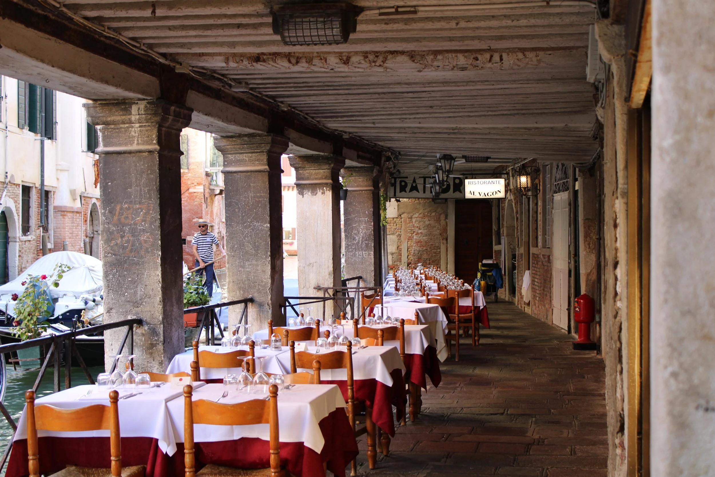 Outdoor restaurant along a canal with tables set for dining under a covered walkway in Venice, Italy, with a man in a striped shirt walking by.
