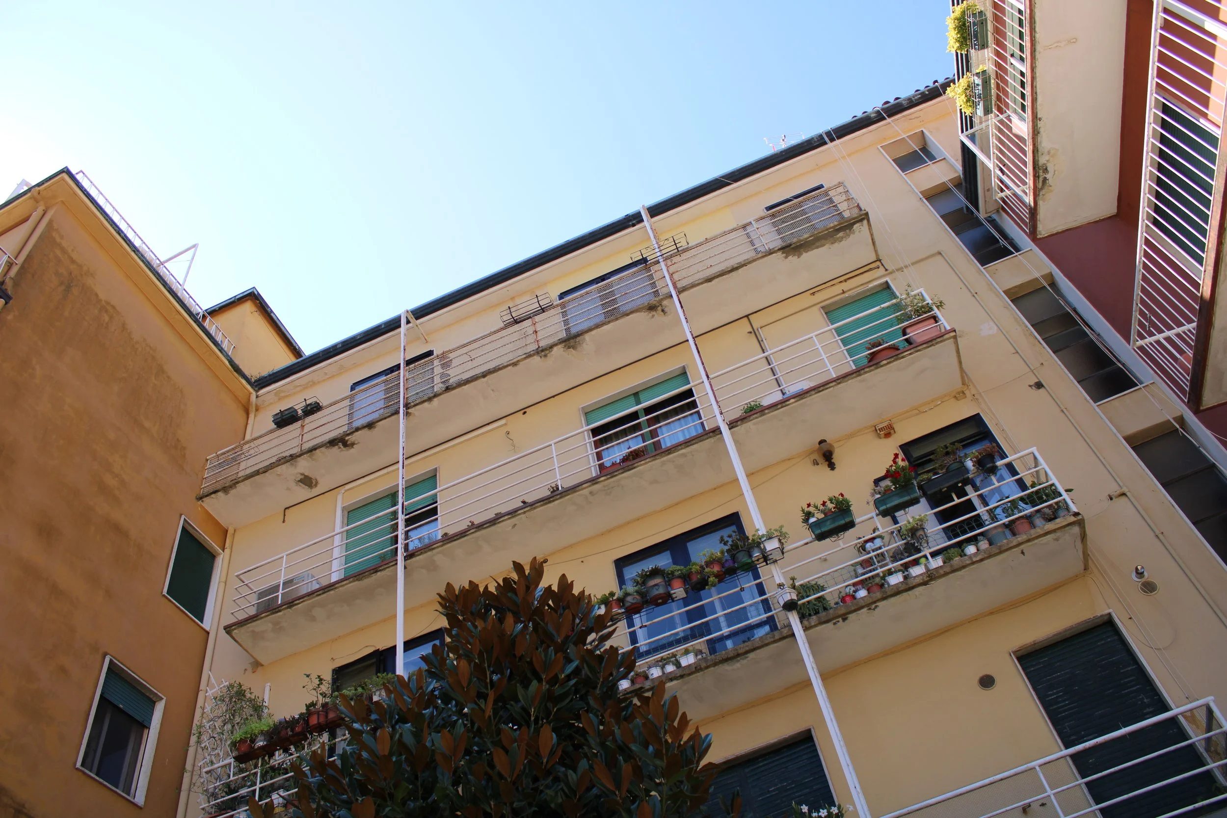 Looking up at a beige apartment building with multiple balconies, some with potted plants and flowers, against a clear blue sky.