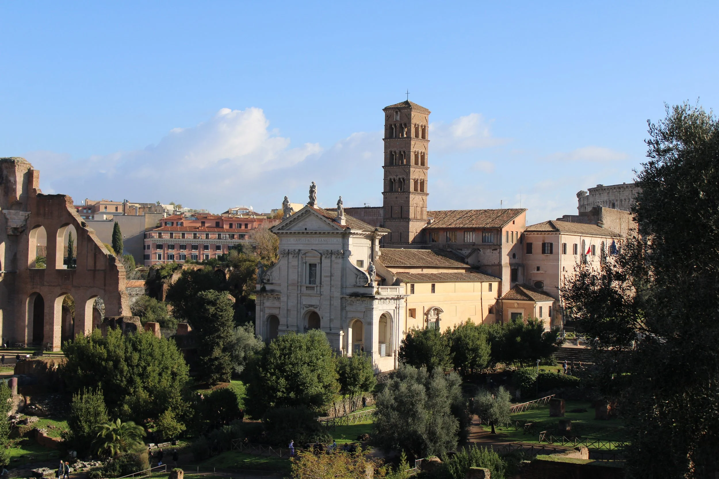 Photo of ancient ruins, a church, and a tall tower in Rome, Italy, with greenery and a blue sky with clouds.