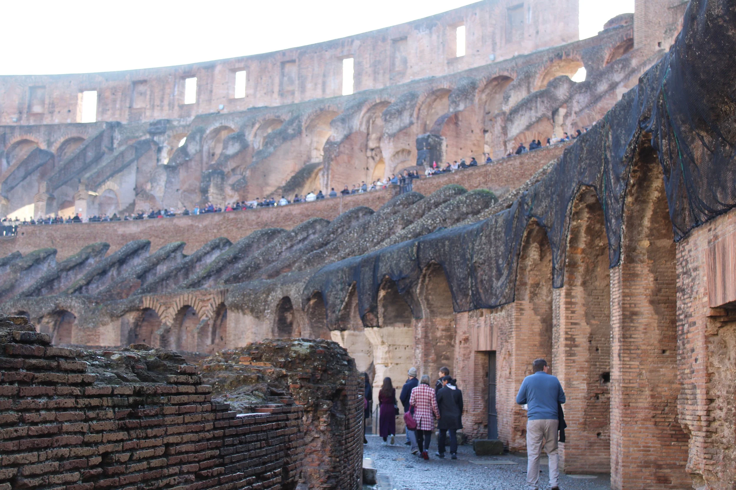 People walking near the base of the historic Roman Colosseum, with the upper tiers and arches visible in the background.