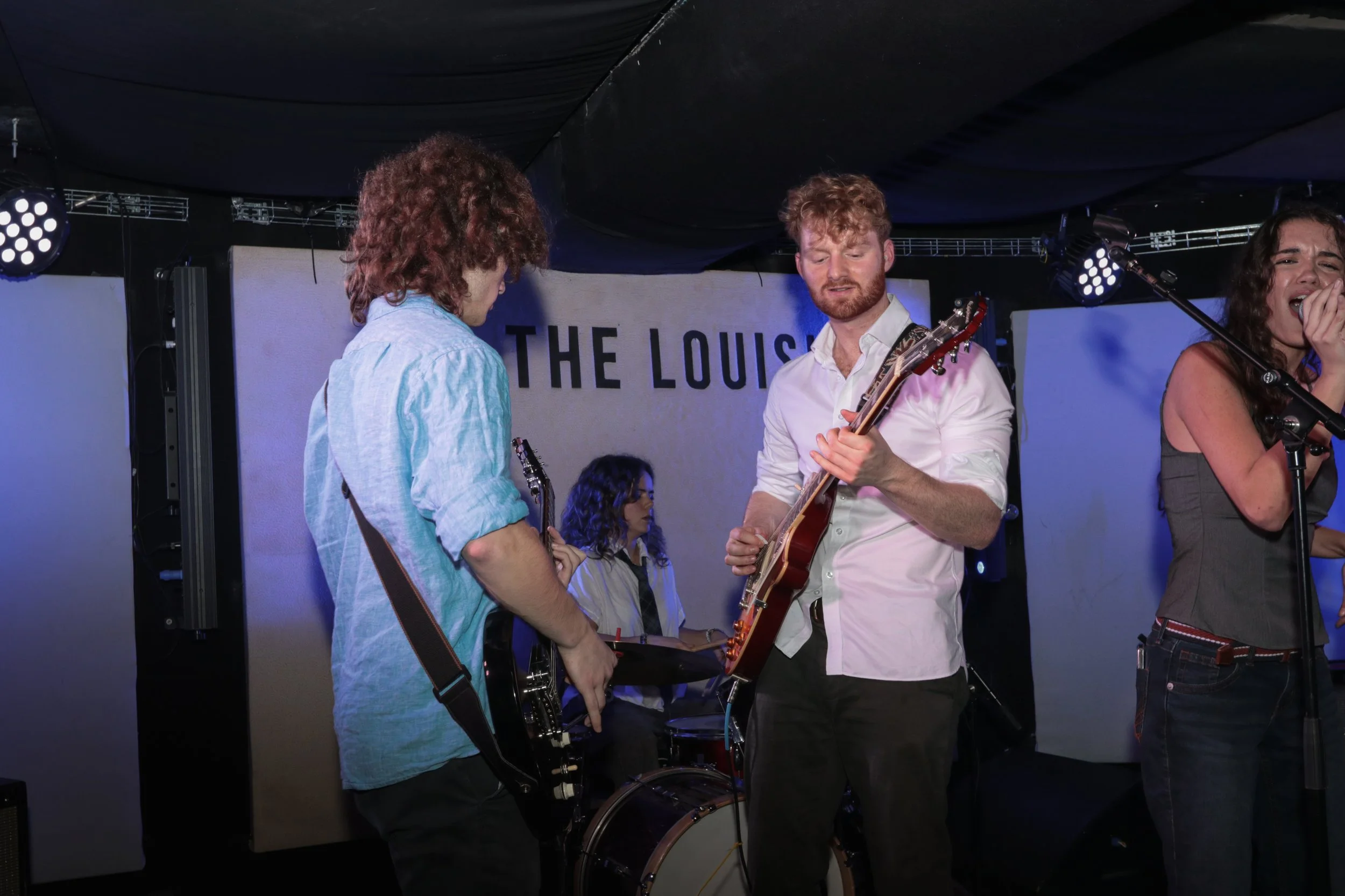 Four musicians performing on stage: a guitarist, a bassist, a drummer, and a vocalist, with a sign reading 'The Loui...' in the background.
