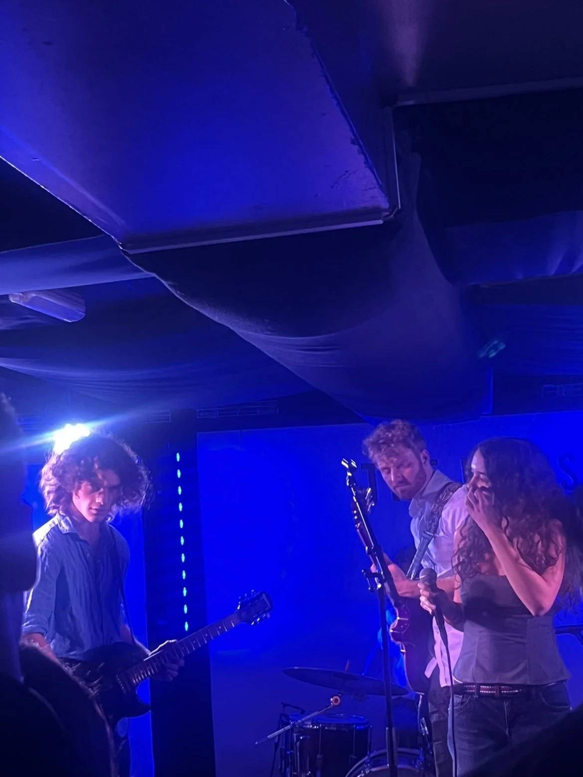 Three musicians perform on stage under blue lighting. One person plays guitar, another stands at a microphone, and a third holds an instrument, with a drum set partly visible.