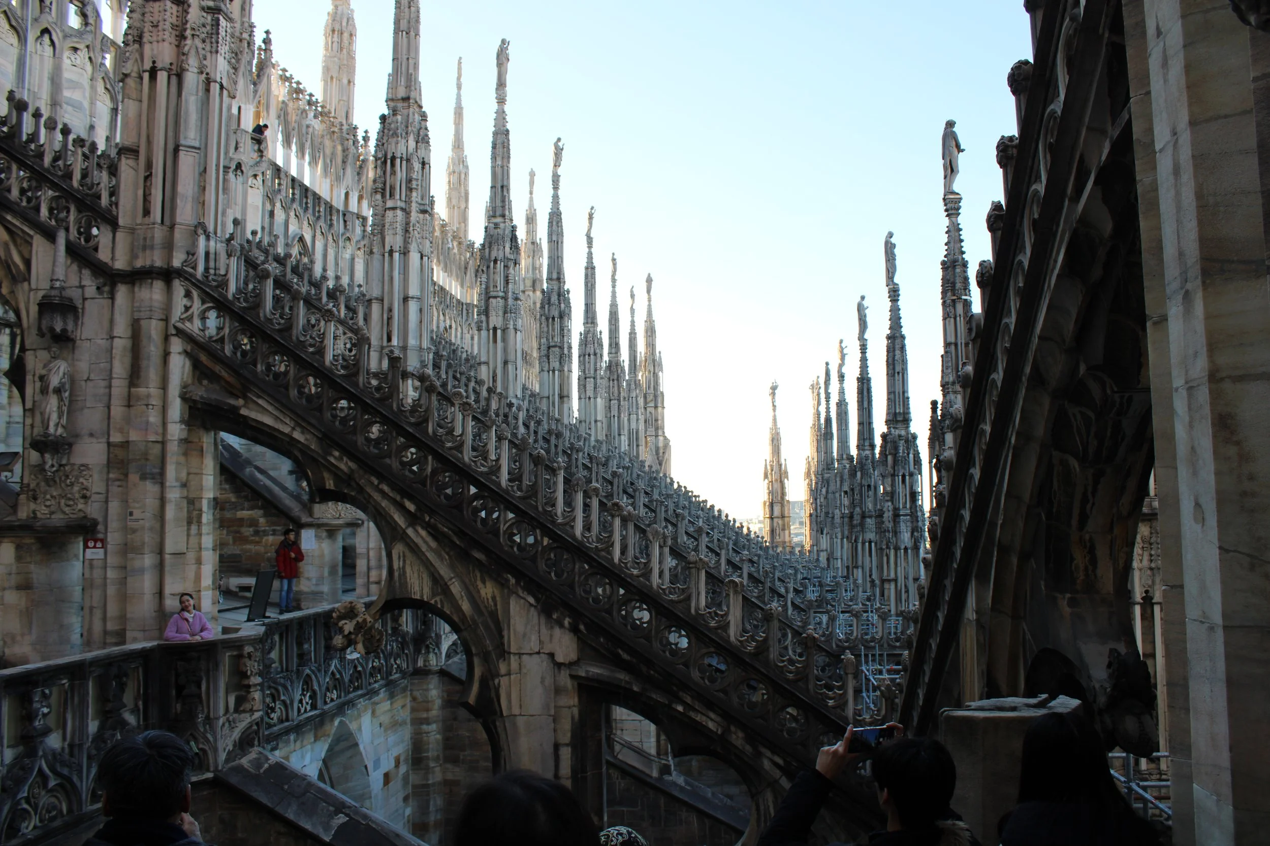 People walking and taking photos outside the Milan Cathedral's intricate Gothic architecture with numerous spires and statues.